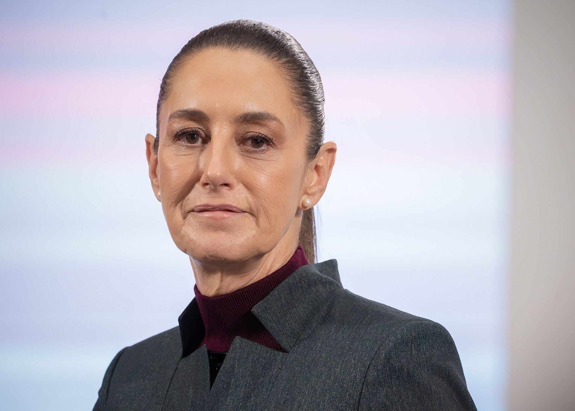 Claudia Sheinbaum, con saco gris y cuello alto vino, mira al frente con un fondo de tonos pastel desenfocado.