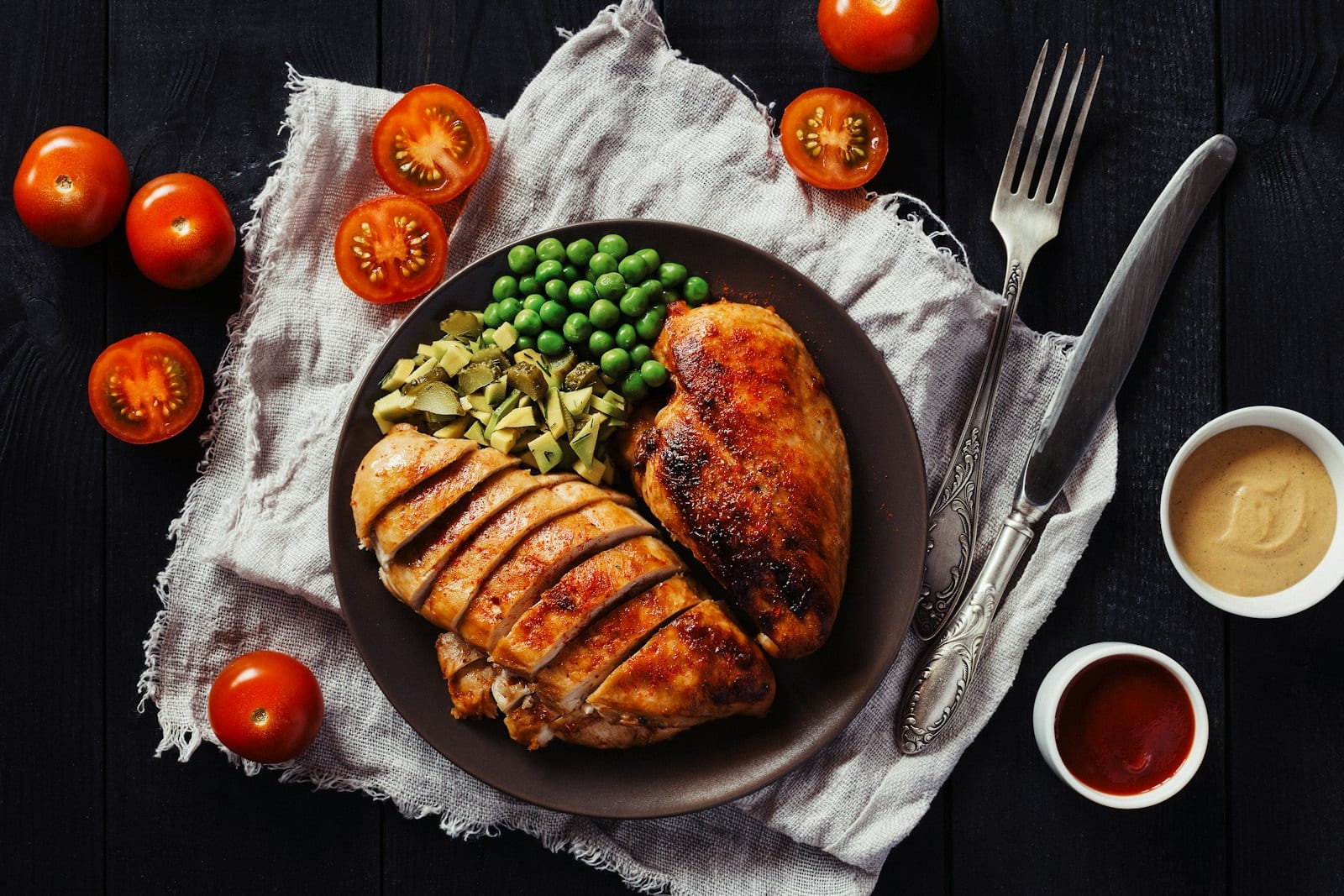 Pechuga de pollo asada y rebanada servida junto a chícharos, pepinillos y jitomates.
