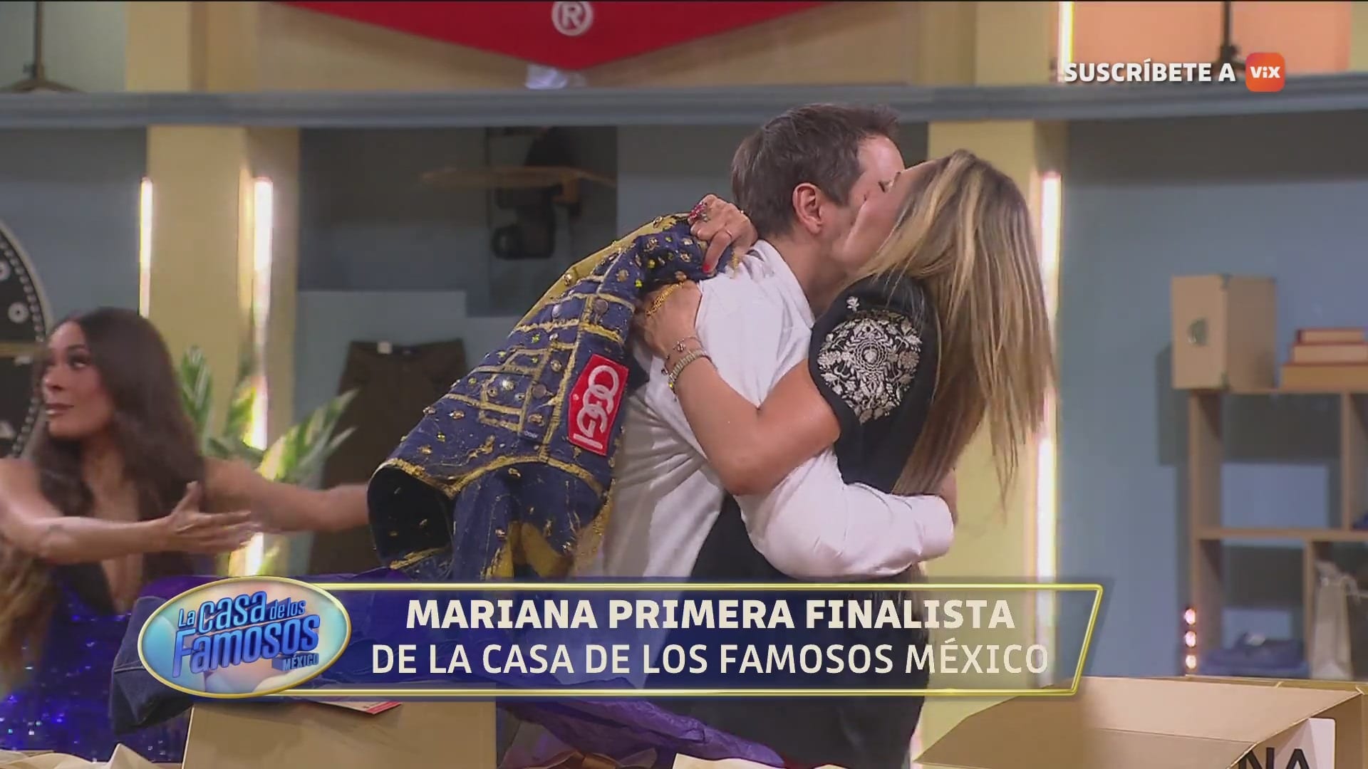 Dos concursantes se abrazan dentro de la casa, con un letrero en pantalla que anuncia a la primera finalista.