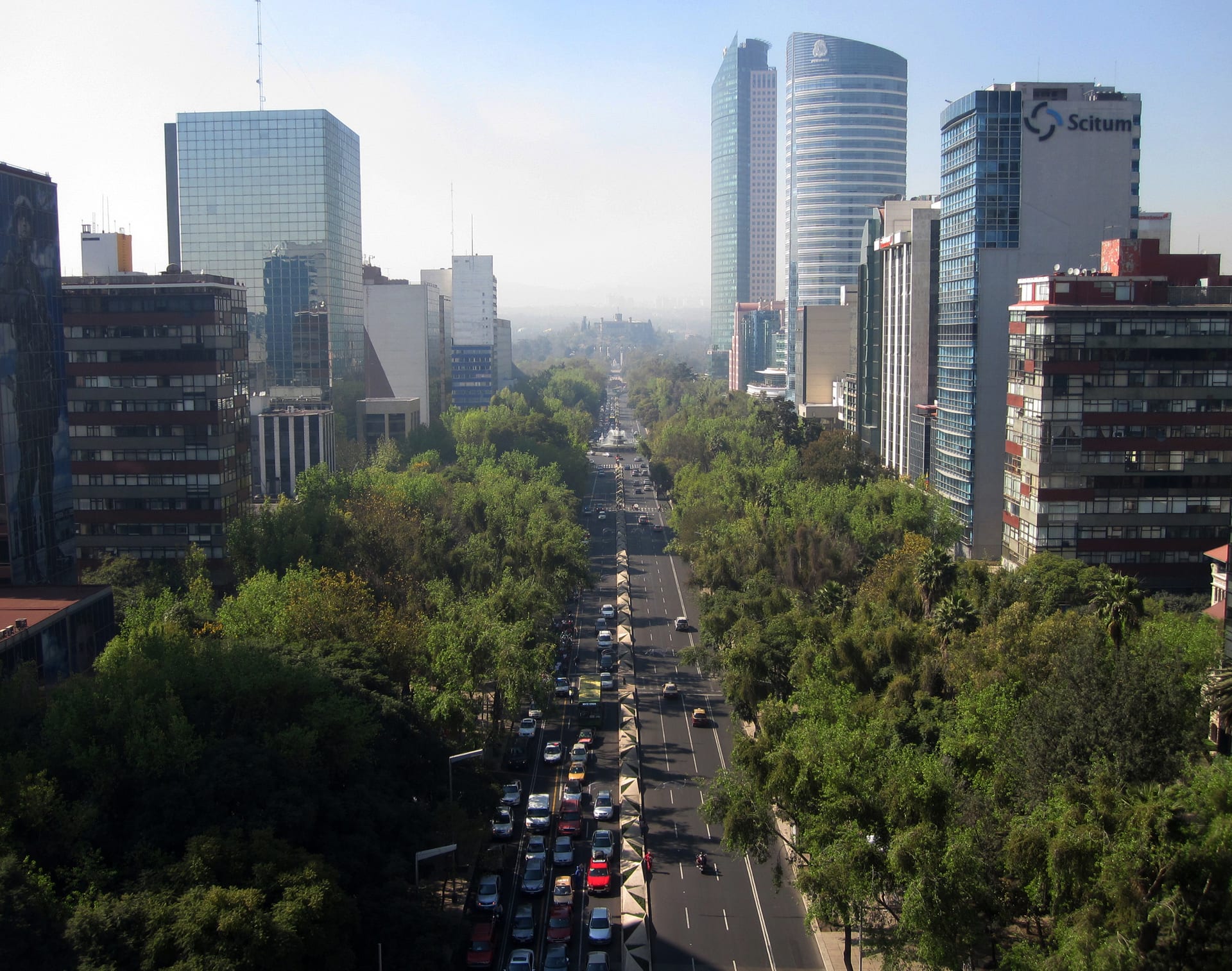 Vista aérea de Paseo de la Reforma con torres de oficinas a los costados, un camellón arbolado y tráfico intenso al centro.
