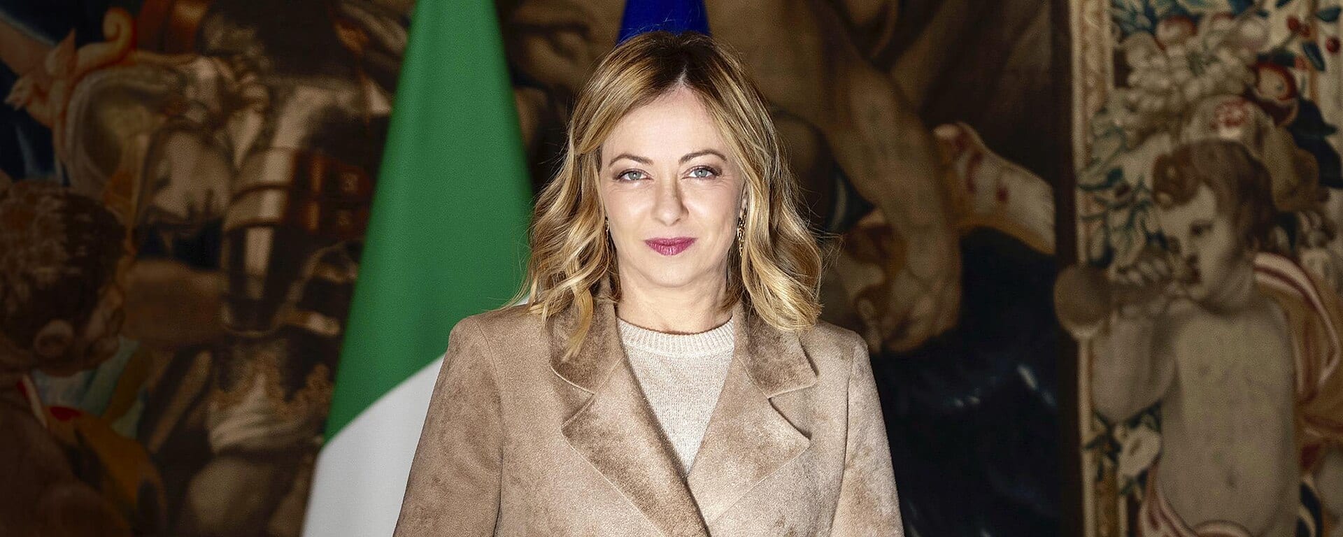 Giorgia Meloni posa de pie con abrigo color camel frente a banderas de Italia y la Unión Europea, con tapices antiguos al fondo.