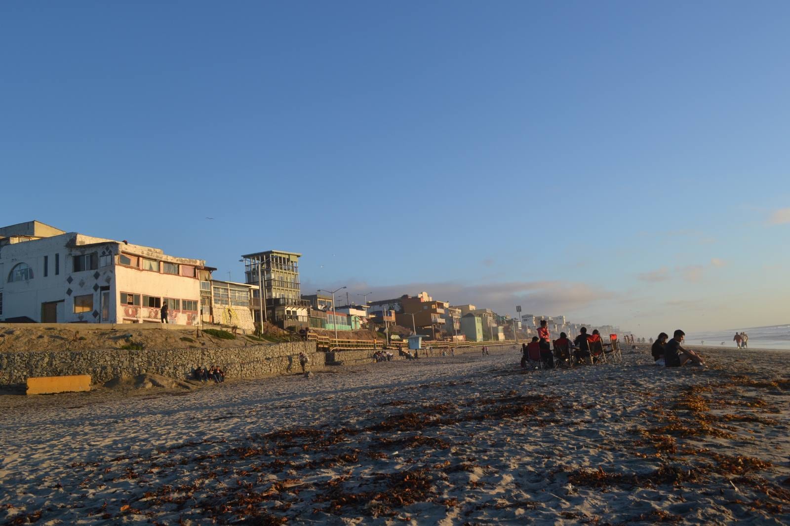 Playa de Playas de Tijuana al atardecer, con edificios frente al mar y grupos de personas sentadas en la arena.