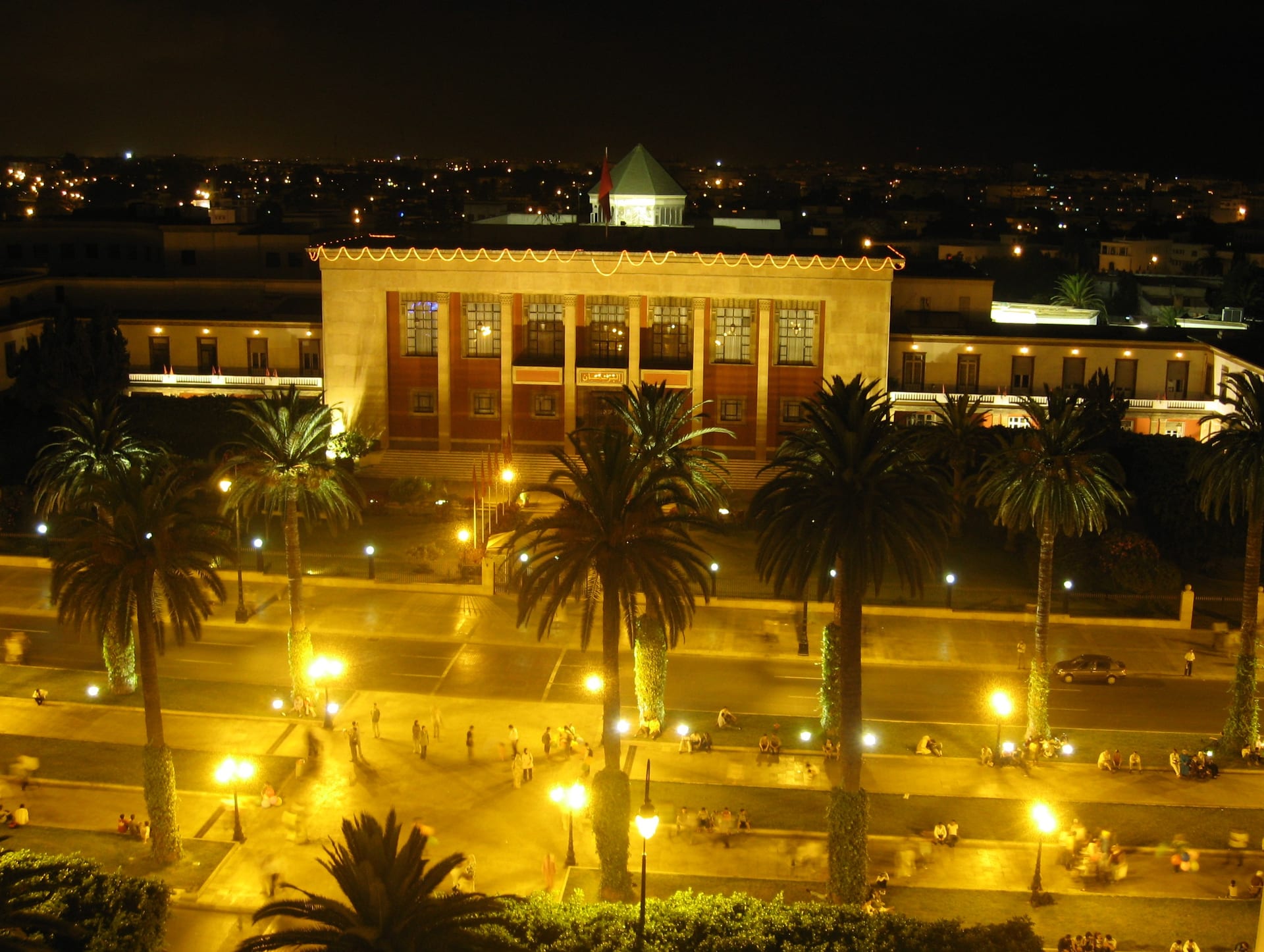 Vista nocturna del edificio del Parlamento de Marruecos en Rabat, iluminado y rodeado de palmeras, con gente reunida en la explanada.