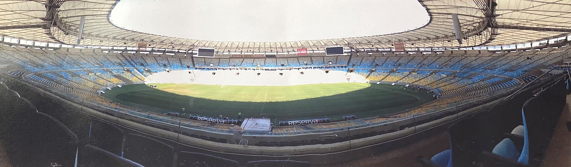 Interior vacío de un estadio con techo blanco curvo, gradas azules y amarillas, y una cancha de pasto verde al centro.