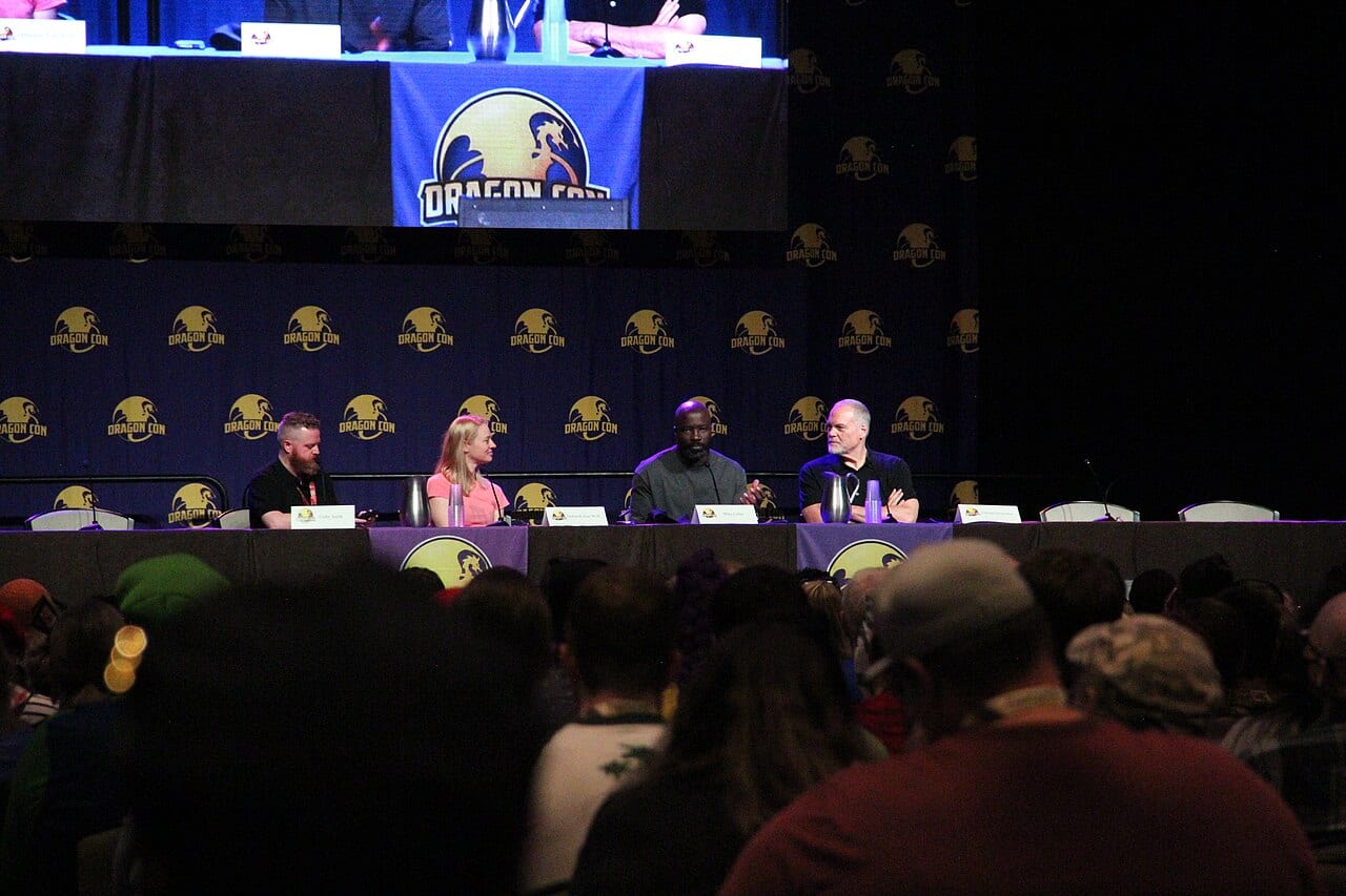 Cuatro personas conversan sentadas en una mesa de panel de convención, con un fondo azul de Dragon Con y público en primer plano.