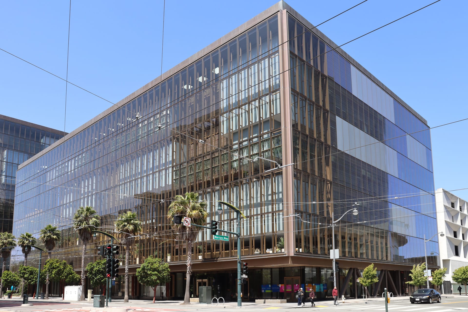 Fachada de vidrio de un edificio de oficinas de varios pisos en una esquina urbana, con palmeras y peatones en la banqueta.
