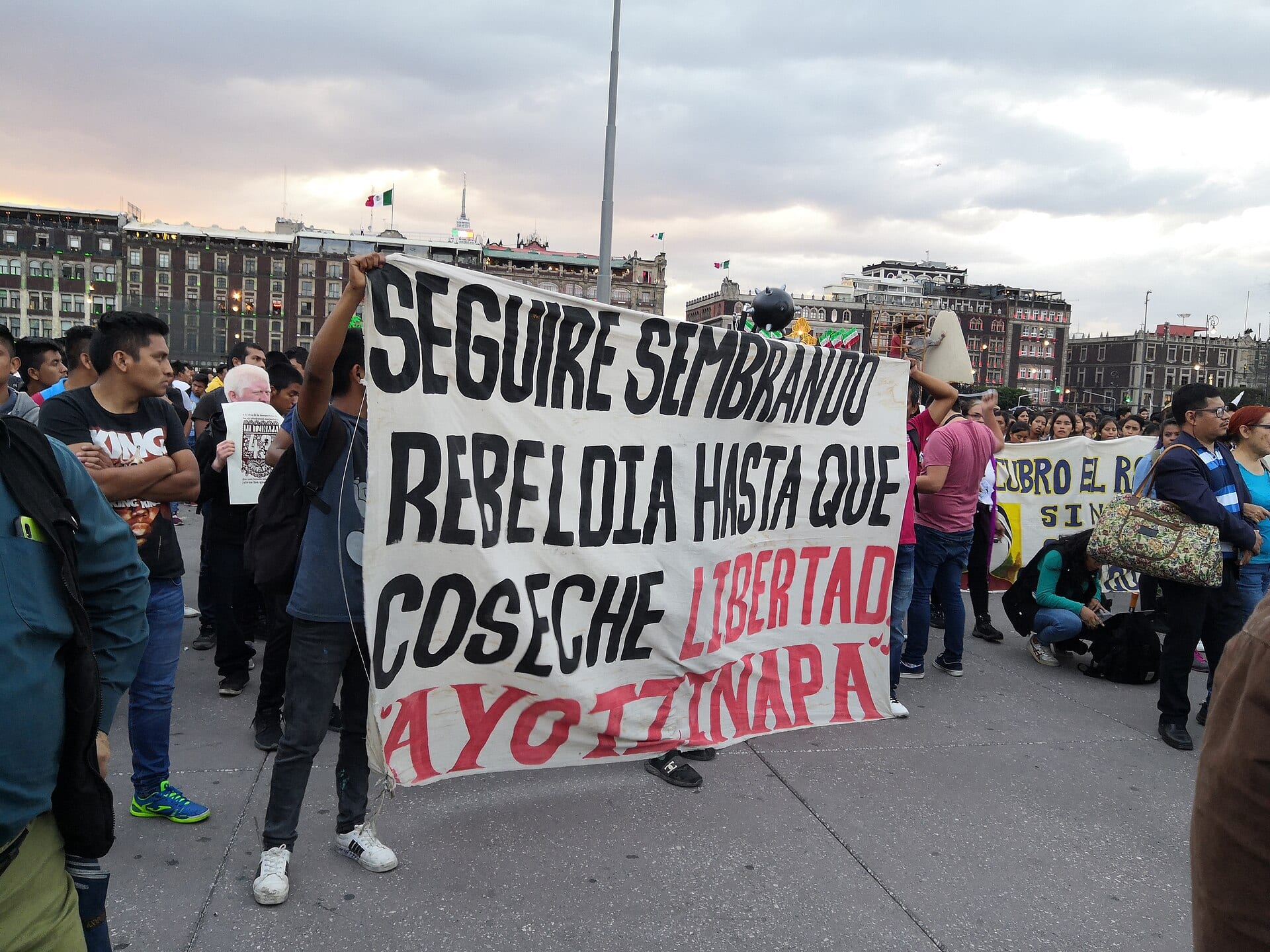Un hombre levanta una manta blanca con la leyenda "Seguiré sembrando rebeldía hasta que coseche libertad, Ayotzinapa" frente a una multitud en el Zócalo.