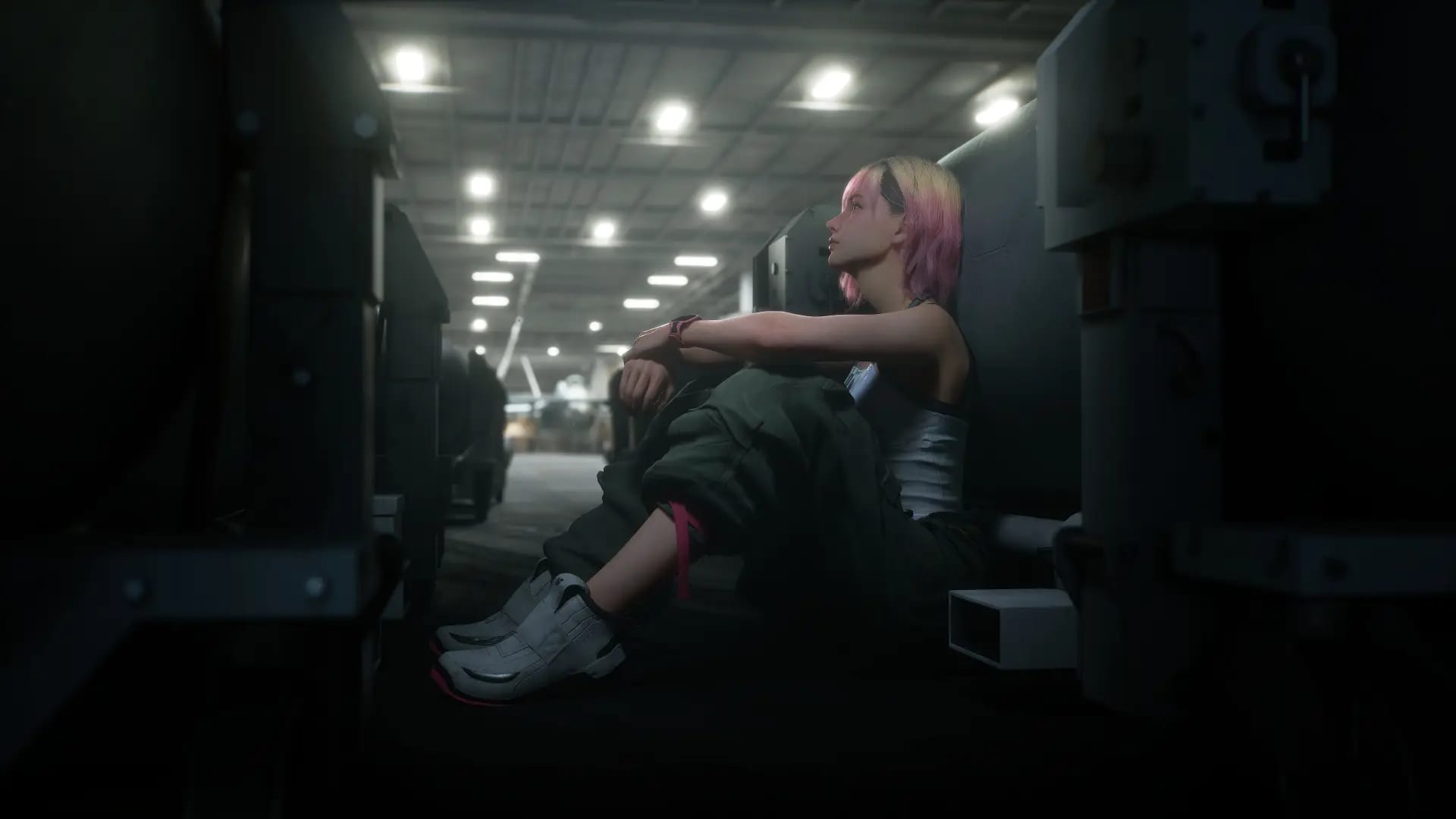 Una joven de cabello rosado y rubio sentada en el piso de un hangar militar en penumbra, rodeada de vehículos y contenedores.