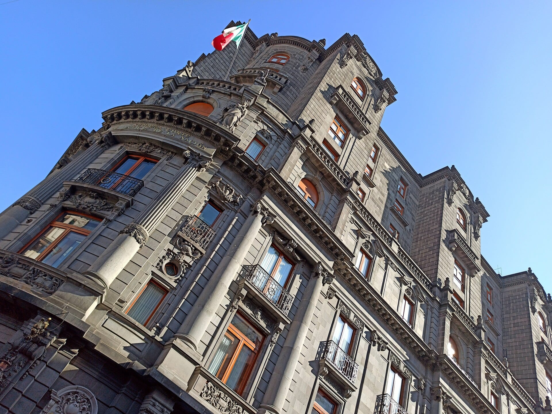 Fachada en piedra gris del edificio de la Suprema Corte vista desde abajo, con la bandera de México ondeando en la torre.