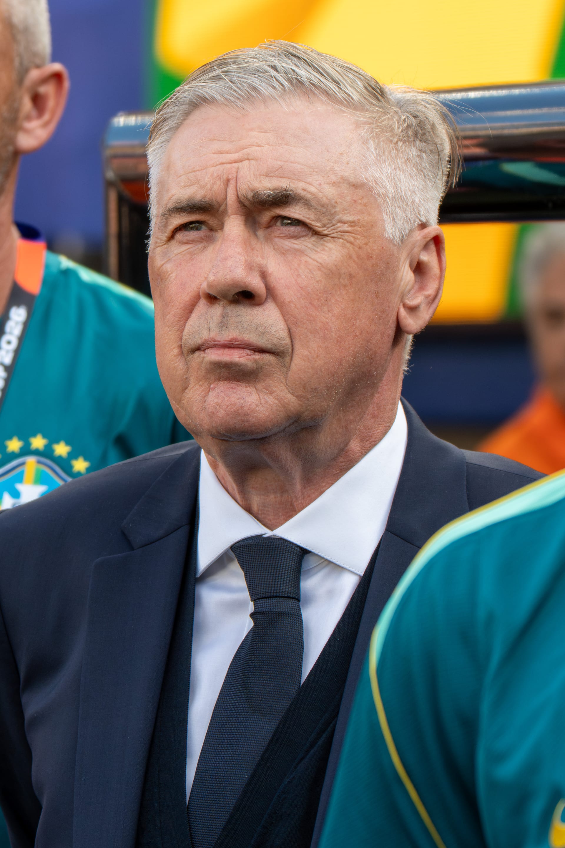 Retrato de Carlo Ancelotti con traje y corbata oscuros, gesto serio, junto a jugadores con playera verde de la selección de Brasil.