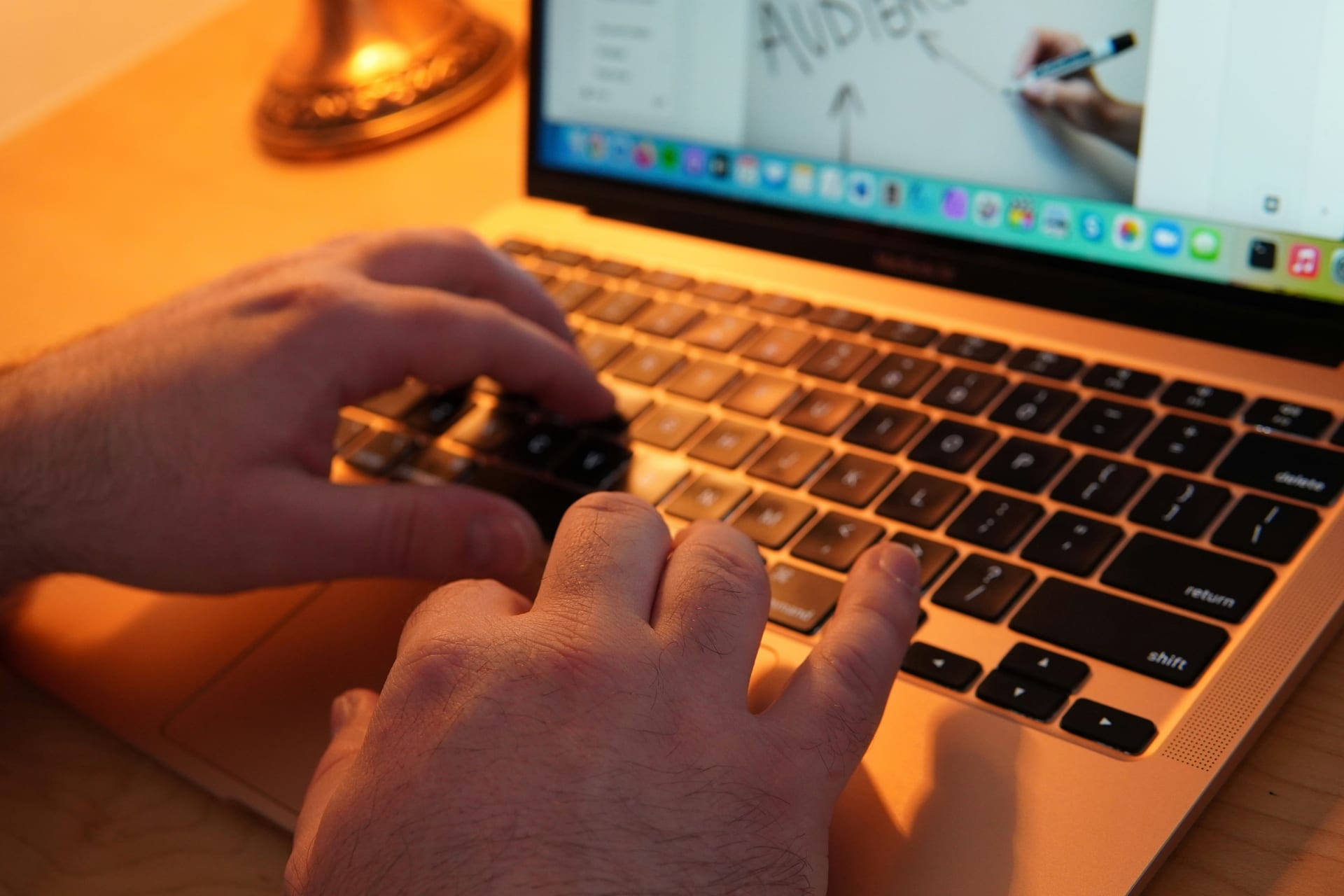 Un par de manos escribe sobre el teclado de una laptop, con la pantalla desenfocada al fondo.