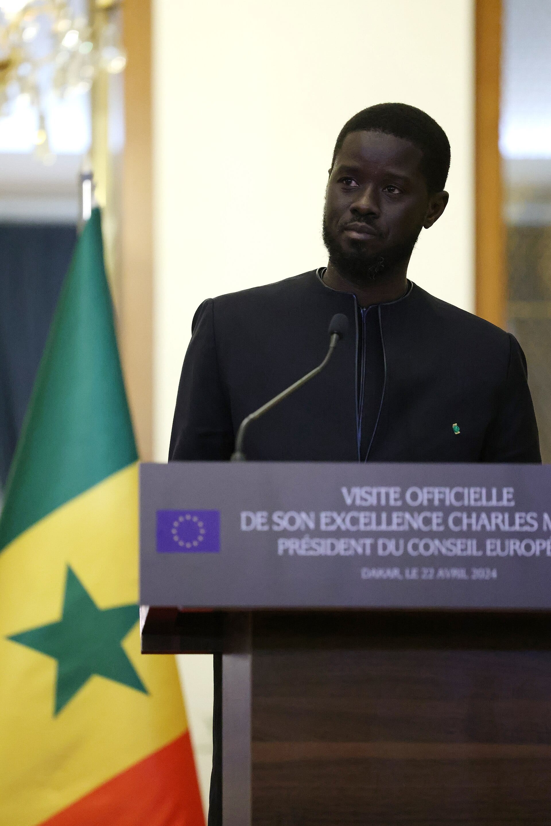 Un hombre con traje oscuro habla frente a un atril con micrófonos y una bandera de Senegal al fondo.