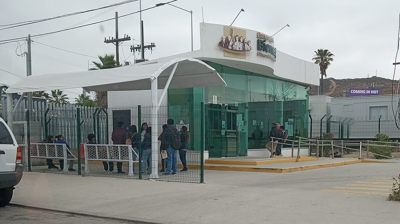 Fachada de una sucursal del Banco del Bienestar, con personas esperando en fila afuera, detrás de una reja, bajo un toldo blanco.