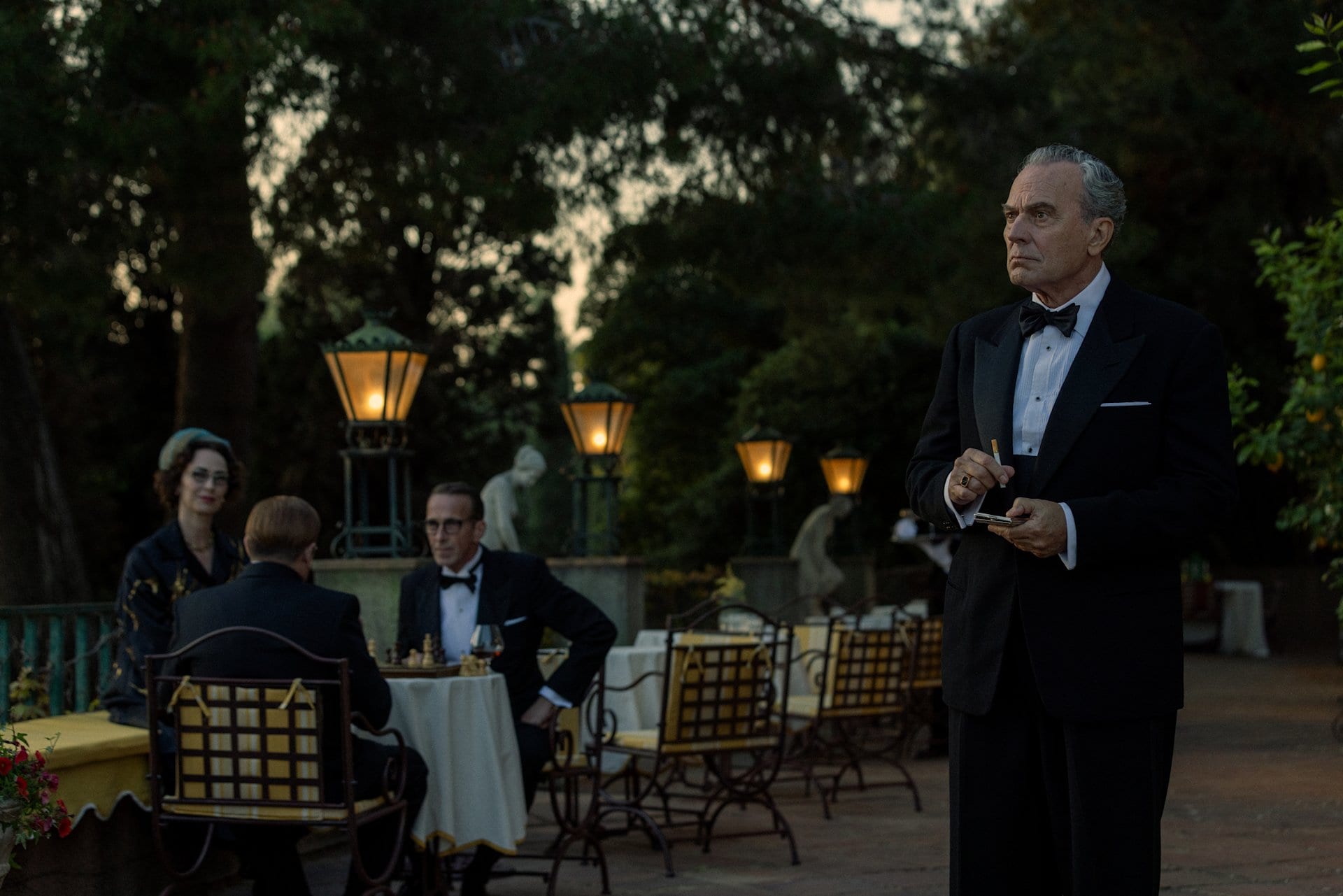 Un actor con esmoquin oscuro fuma de pie en una terraza con faroles encendidos, mientras otros tres huéspedes juegan ajedrez en una mesa detrás de él.