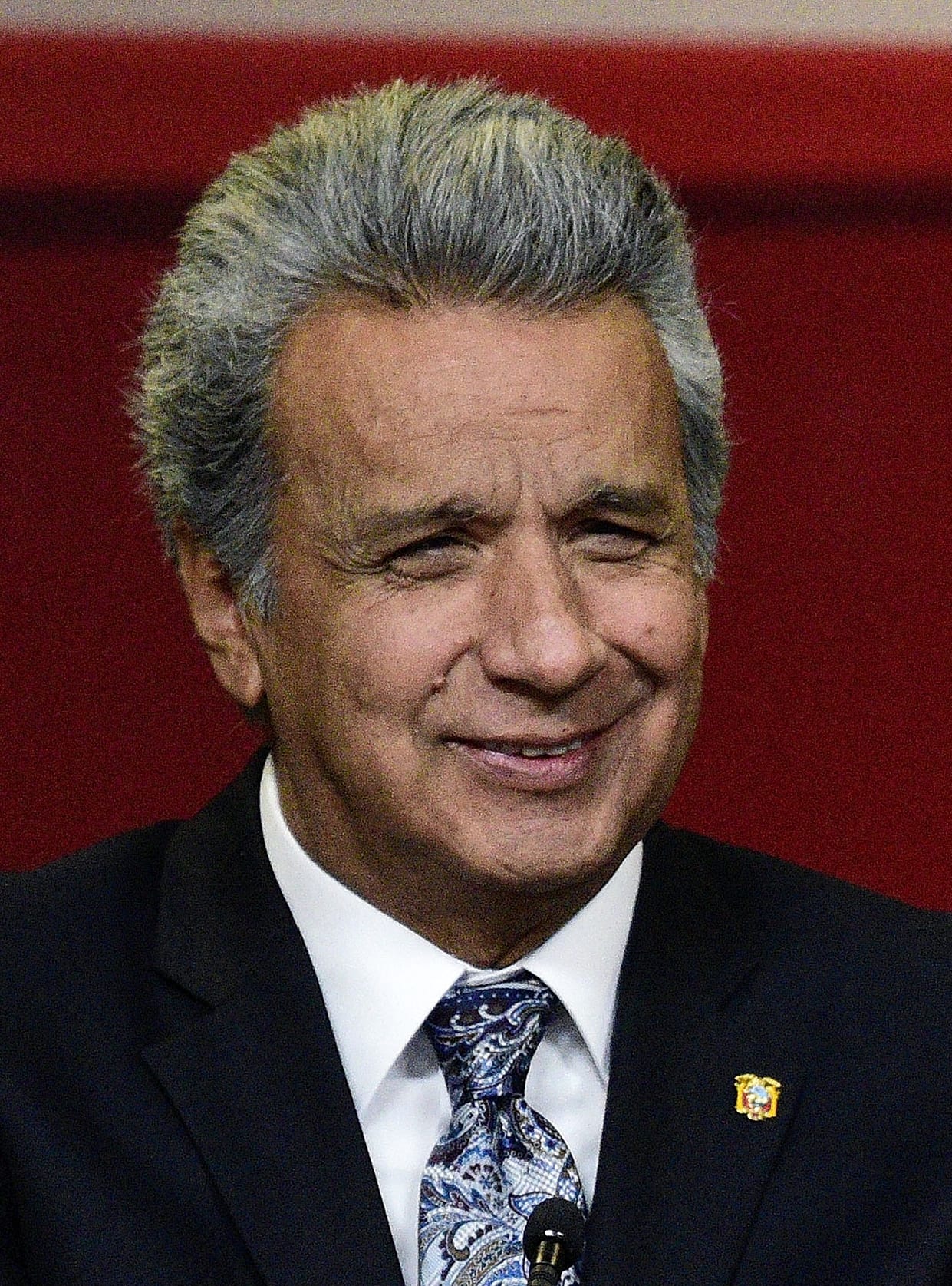 Un hombre de cabello canoso y traje oscuro con corbata azul sonríe de frente ante un fondo rojo.