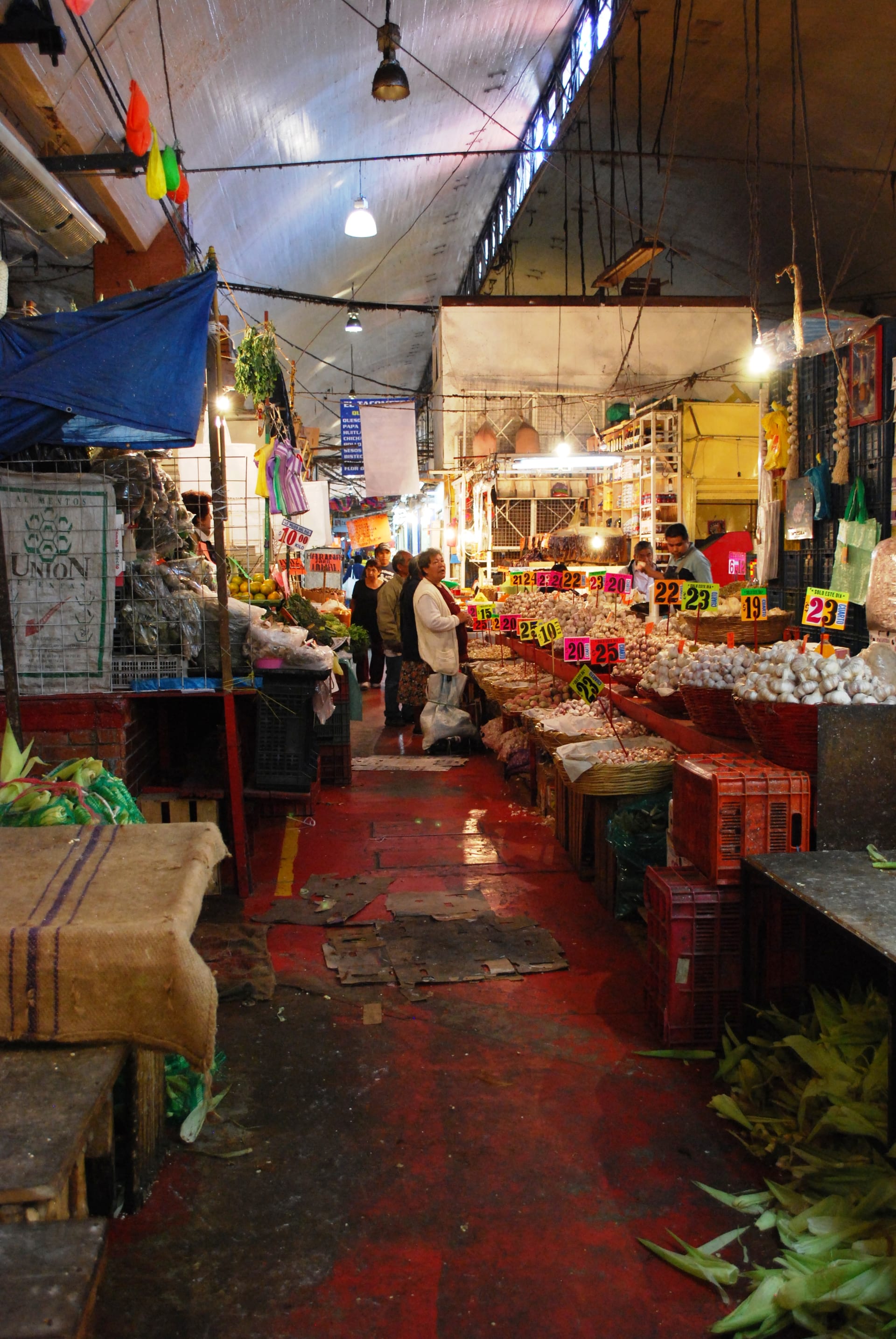 Pasillo cubierto de un mercado con puestos de ajo, letreros de precios de colores y clientes caminando entre los puestos.
