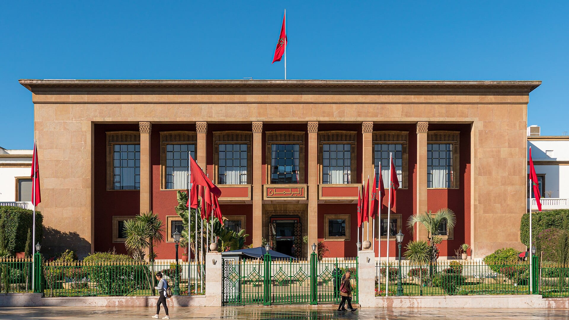 La fachada del Parlamento de Marruecos en Rabat, con banderas rojas ondeando frente a la entrada y dos personas caminando.