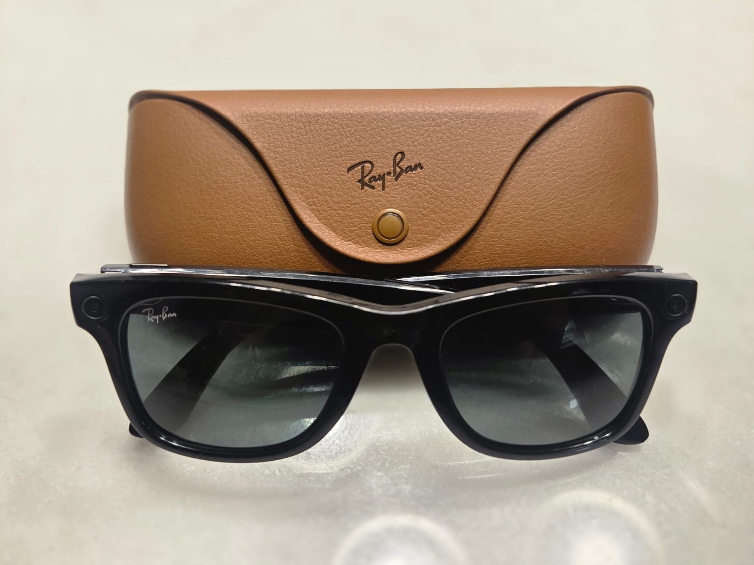 Un par de gafas de sol negras con una cámara diminuta en cada patilla, apoyadas frente a un estuche de piel café con el logotipo Ray-Ban.