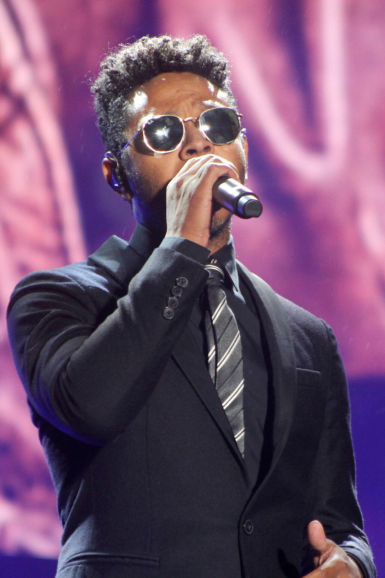 Kalimba canta con lentes de sol y traje oscuro frente a un micrófono, sobre un escenario con luz rosa.