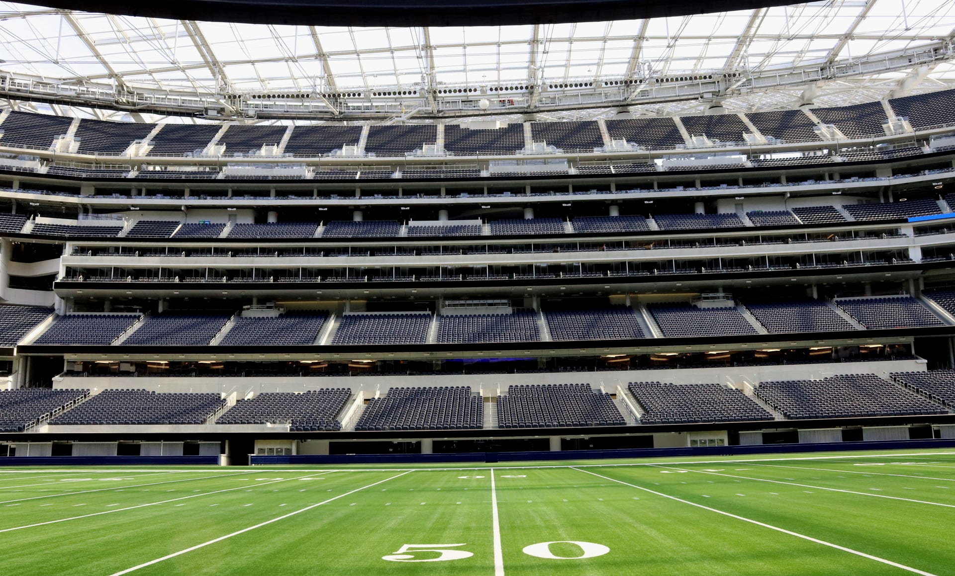Interior de un estadio de futbol americano techado, con las gradas vacías y el campo visto desde la línea de 50 yardas.