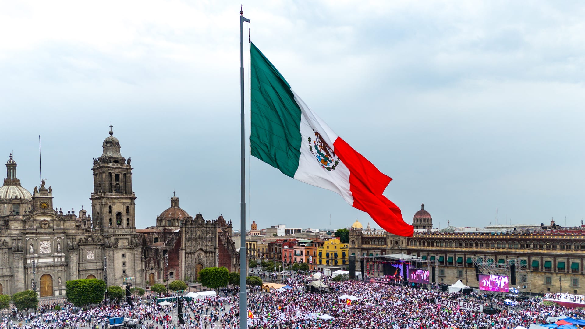 Vista aérea de la bandera monumental de México ondeando sobre una multitud reunida en el Zócalo, con la Catedral Metropolitana al fondo.