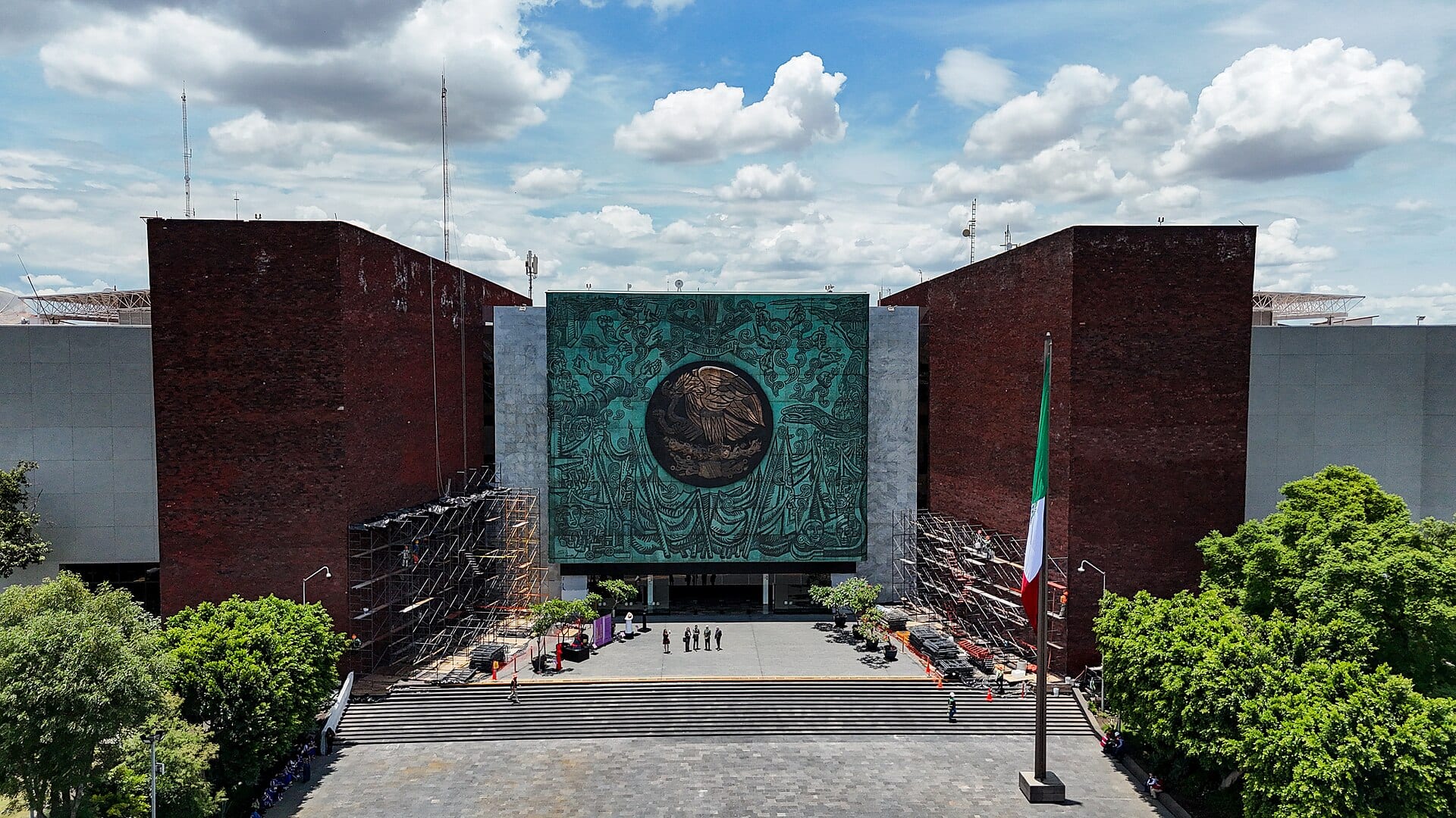 Vista aérea del Palacio Legislativo de San Lázaro, con el escudo nacional en relieve sobre la fachada y una bandera mexicana ondeando junto a la explanada.