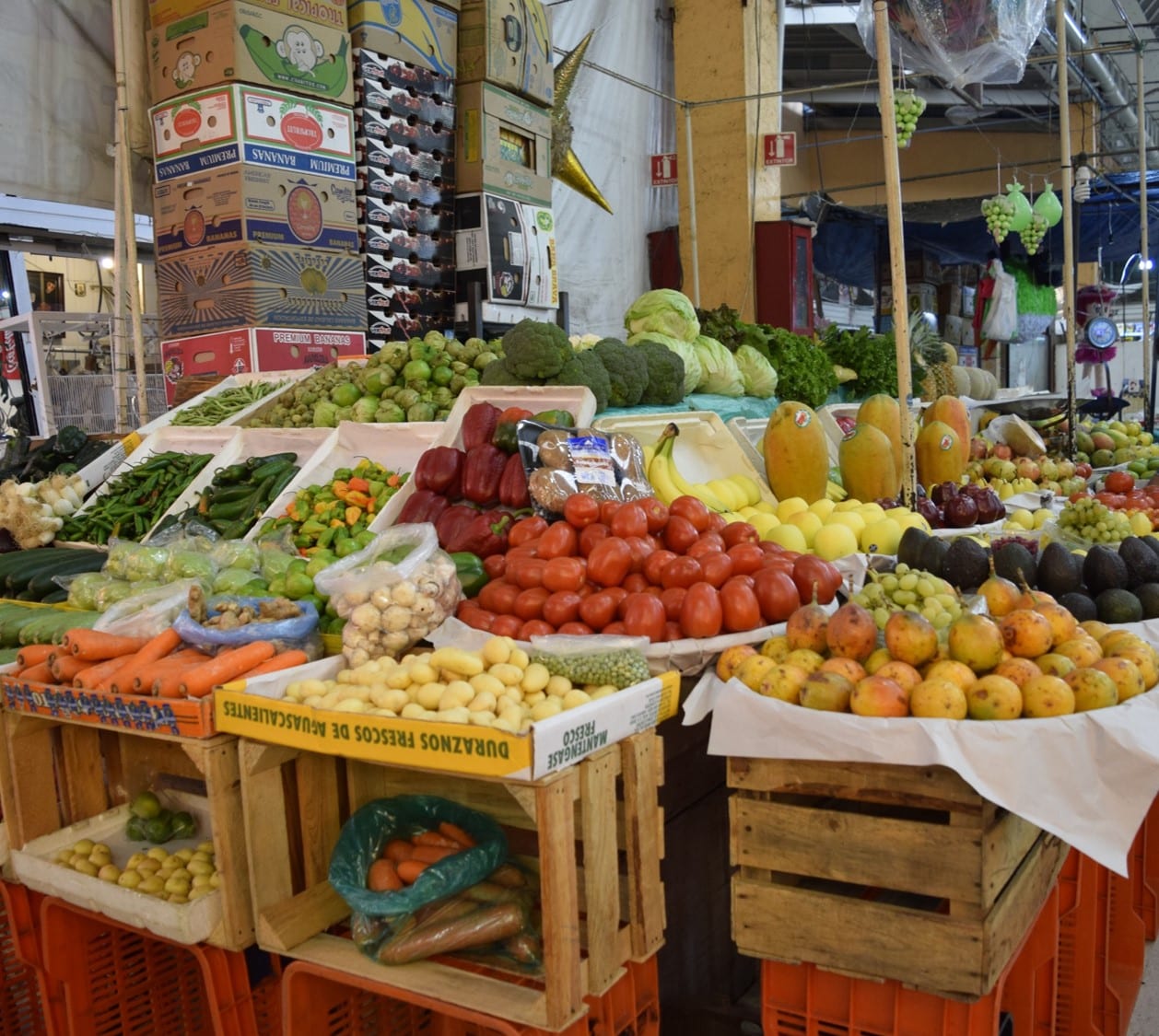 Puesto de mercado con jitomates, plátanos, papayas, brócoli, zanahorias y otras frutas y verduras apiladas.