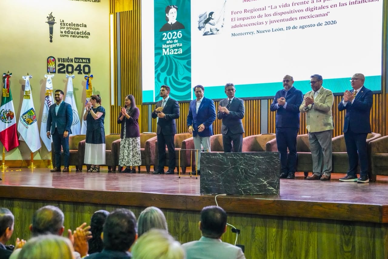 Funcionarios aplauden de pie sobre un escenario, con una pantalla detrás que anuncia el foro "La vida frente a la pantalla" y asistentes de espaldas en primer plano.