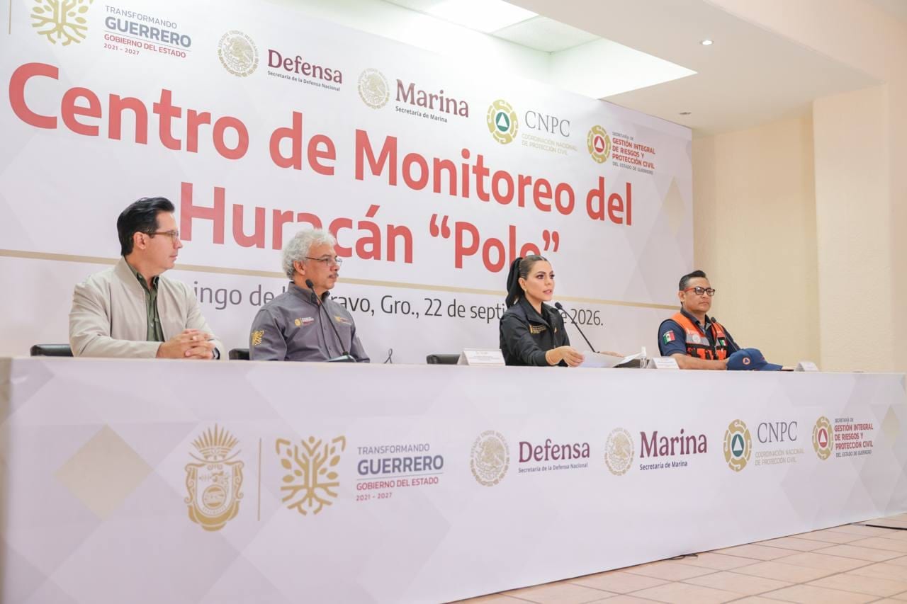 Cuatro personas sentadas en una mesa de conferencia, una mujer habla ante micrófonos frente a un fondo que anuncia un centro de monitoreo del ciclón.