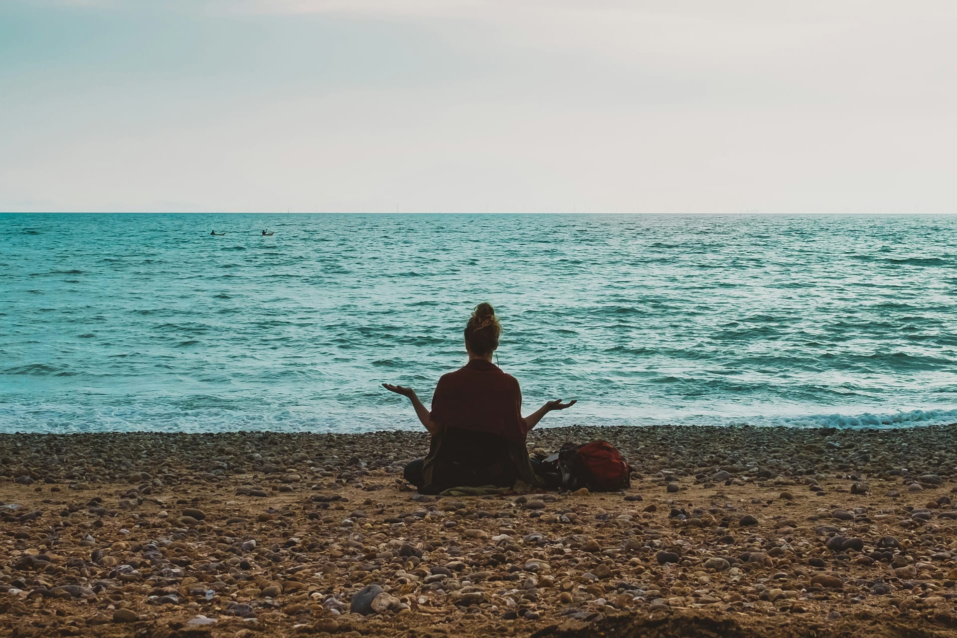 Una persona sentada sobre piedras de playa hace un gesto de meditación frente al mar.
