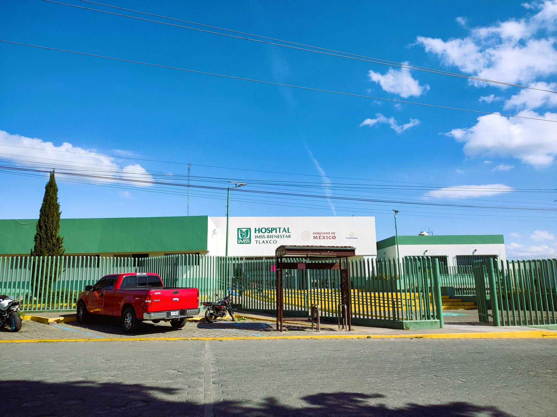 Fachada blanca y verde del Hospital IMSS-Bienestar de Tlaxco, con una reja perimetral y una camioneta roja estacionada en la entrada.