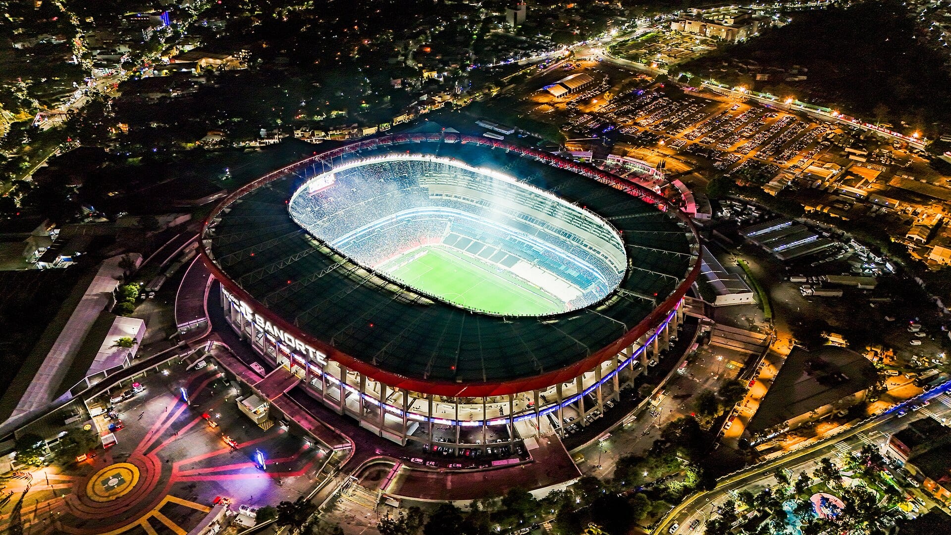 Vista aérea nocturna de un estadio de futbol lleno y techado durante un partido, con el nombre Banorte iluminado en la fachada.
