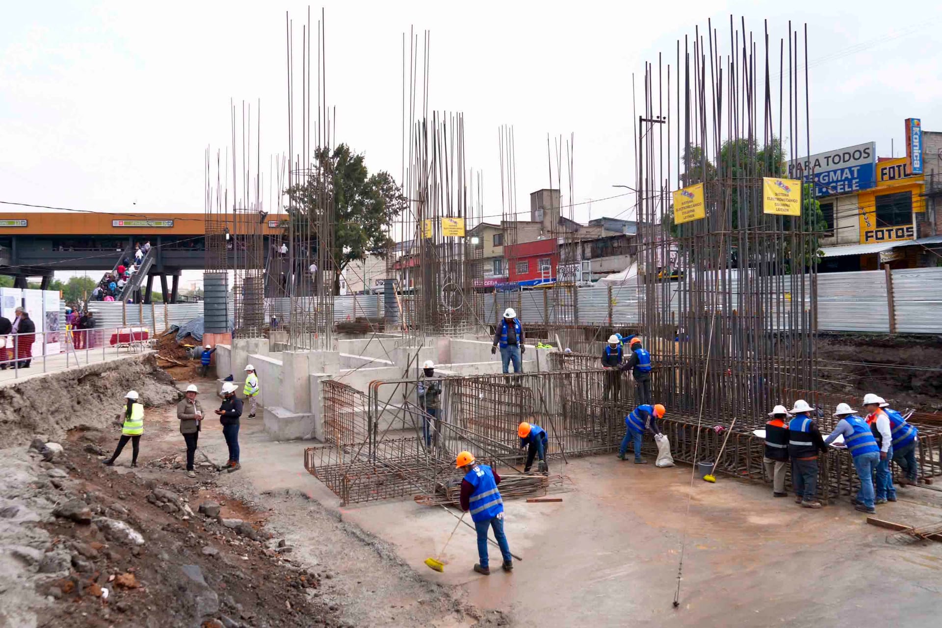 Trabajadores con casco y chaleco arman varillas y cimbra de concreto en la obra de una estación de Cablebús, junto a una estación elevada de transporte.