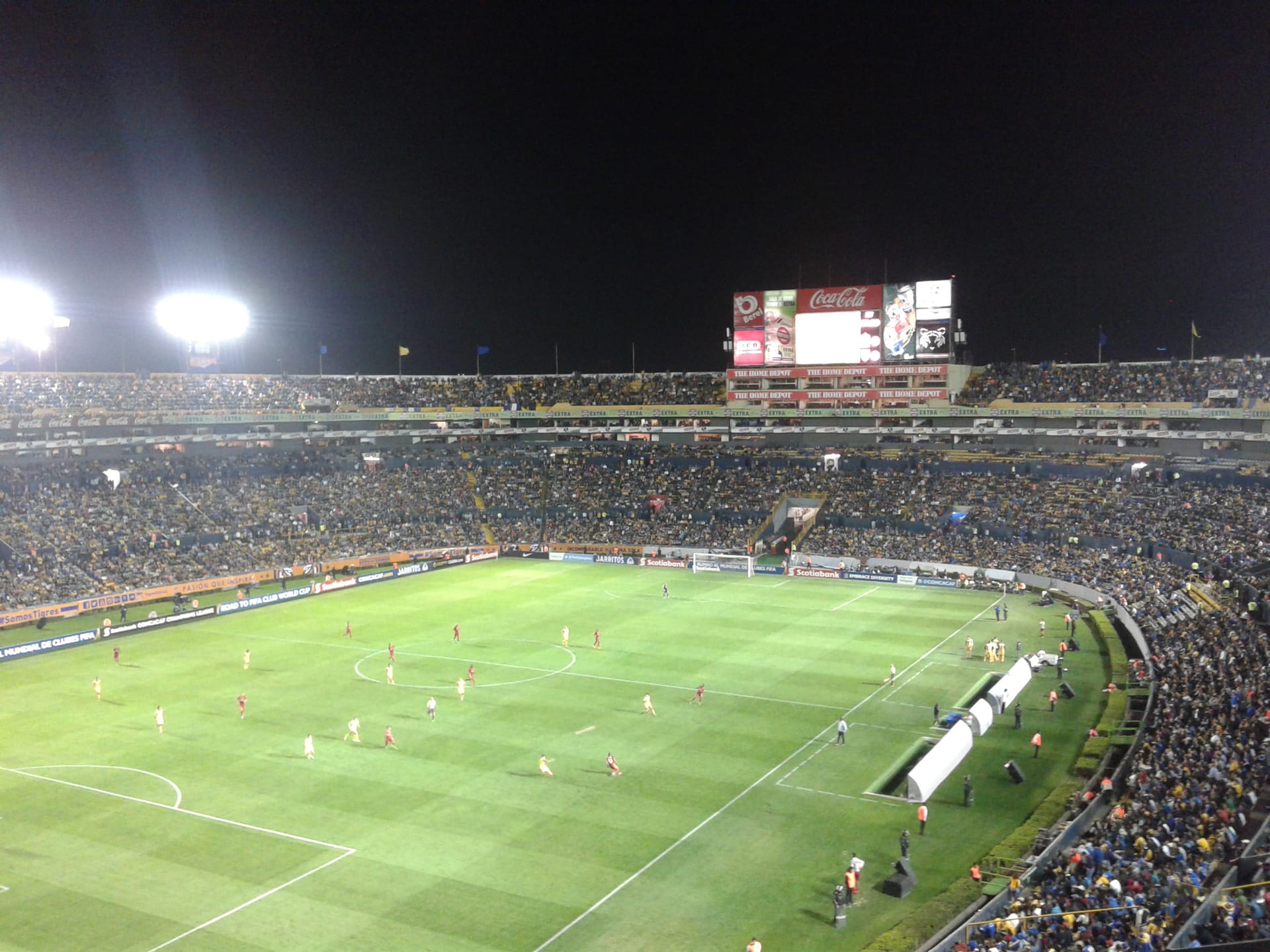 Un estadio de futbol lleno visto desde las gradas altas durante un partido nocturno, con jugadores en la cancha iluminada.