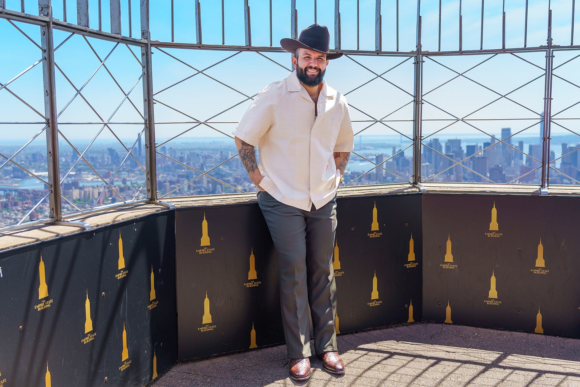 Carín León, con sombrero negro y camisa clara, sonríe con el perfil de Nueva York de fondo desde un mirador.
