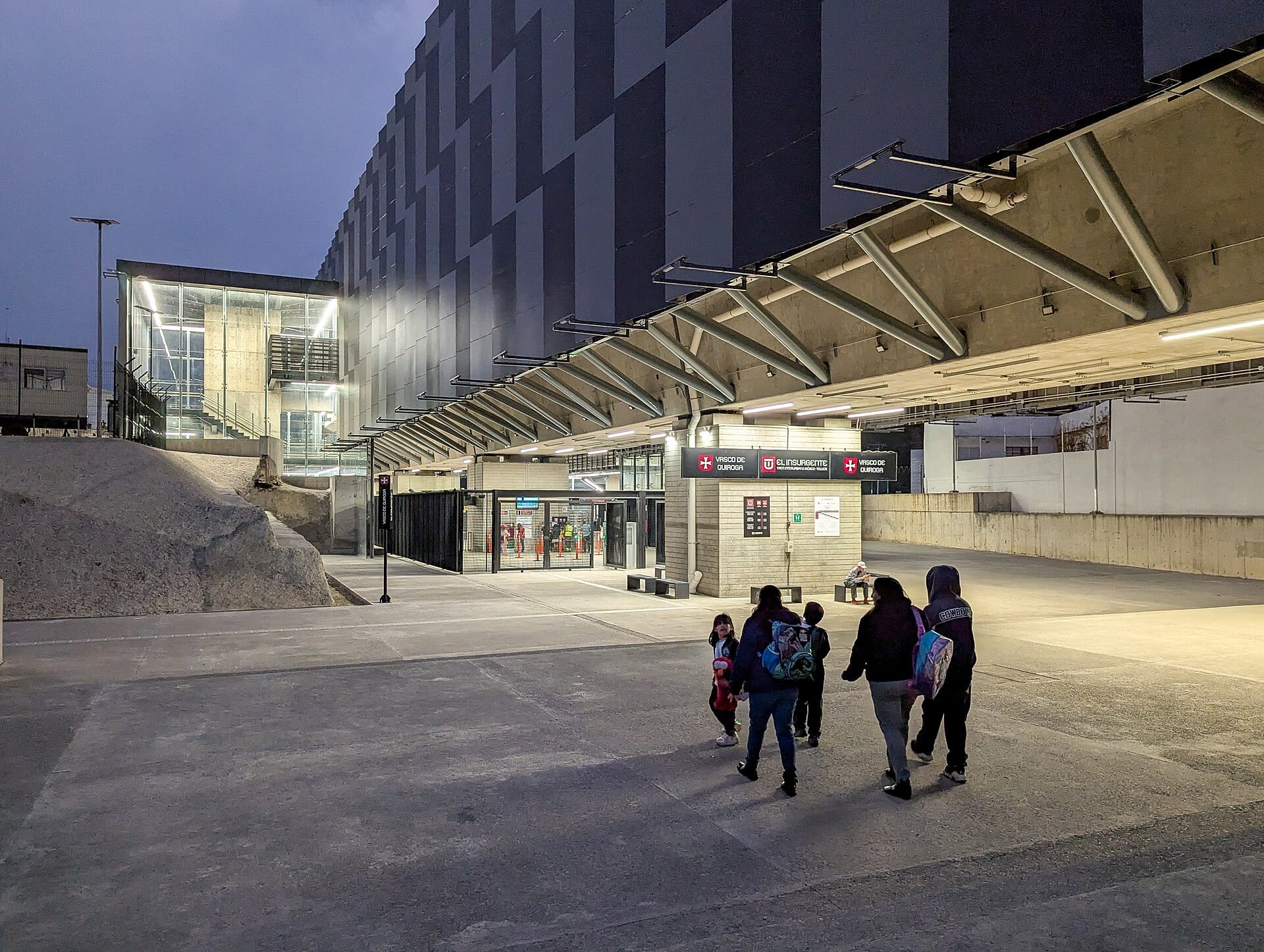 Entrada iluminada de noche a la estación Vasco de Quiroga del Tren El Insurgente, con un grupo de pasajeros caminando hacia el acceso.