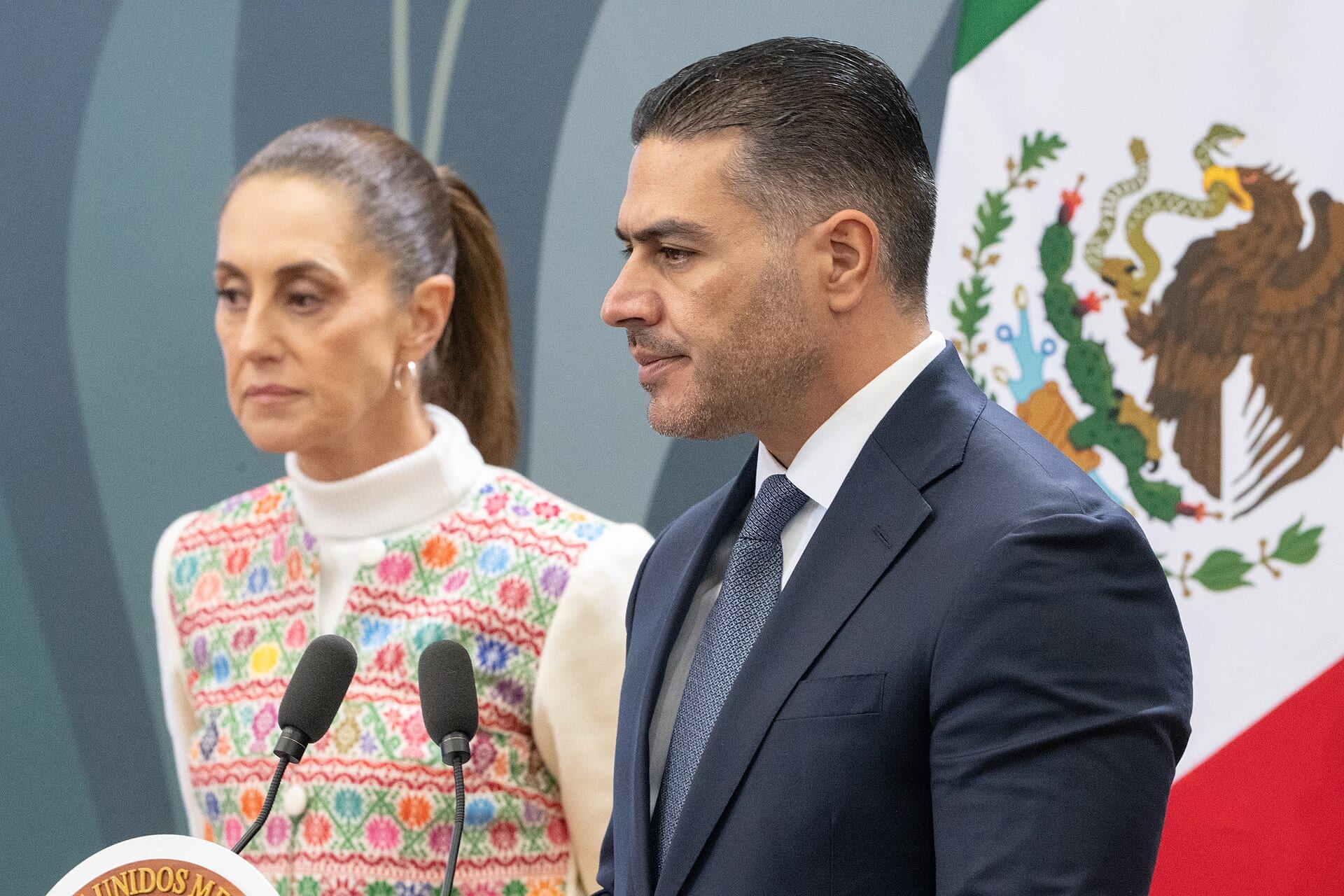 Omar García Harfuch, de traje azul marino, junto a la presidenta Claudia Sheinbaum ante el podio presidencial durante una conferencia matutina.