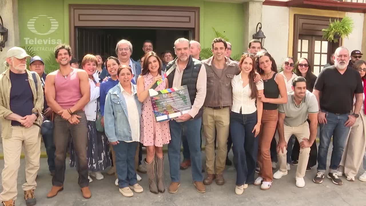 Un grupo numeroso de actores y actrices posa sonriente afuera de un foro, mientras dos de ellos sostienen una claqueta de producción.