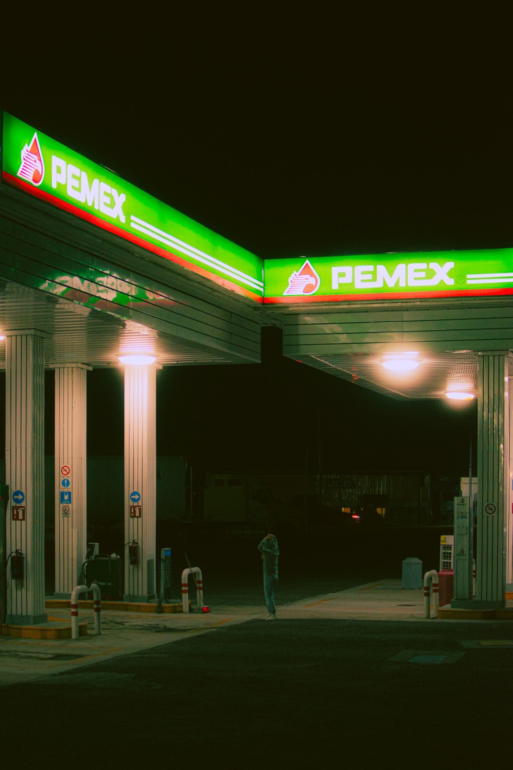 Una figura solitaria camina de noche junto a los surtidores iluminados de una gasolinera Pemex.