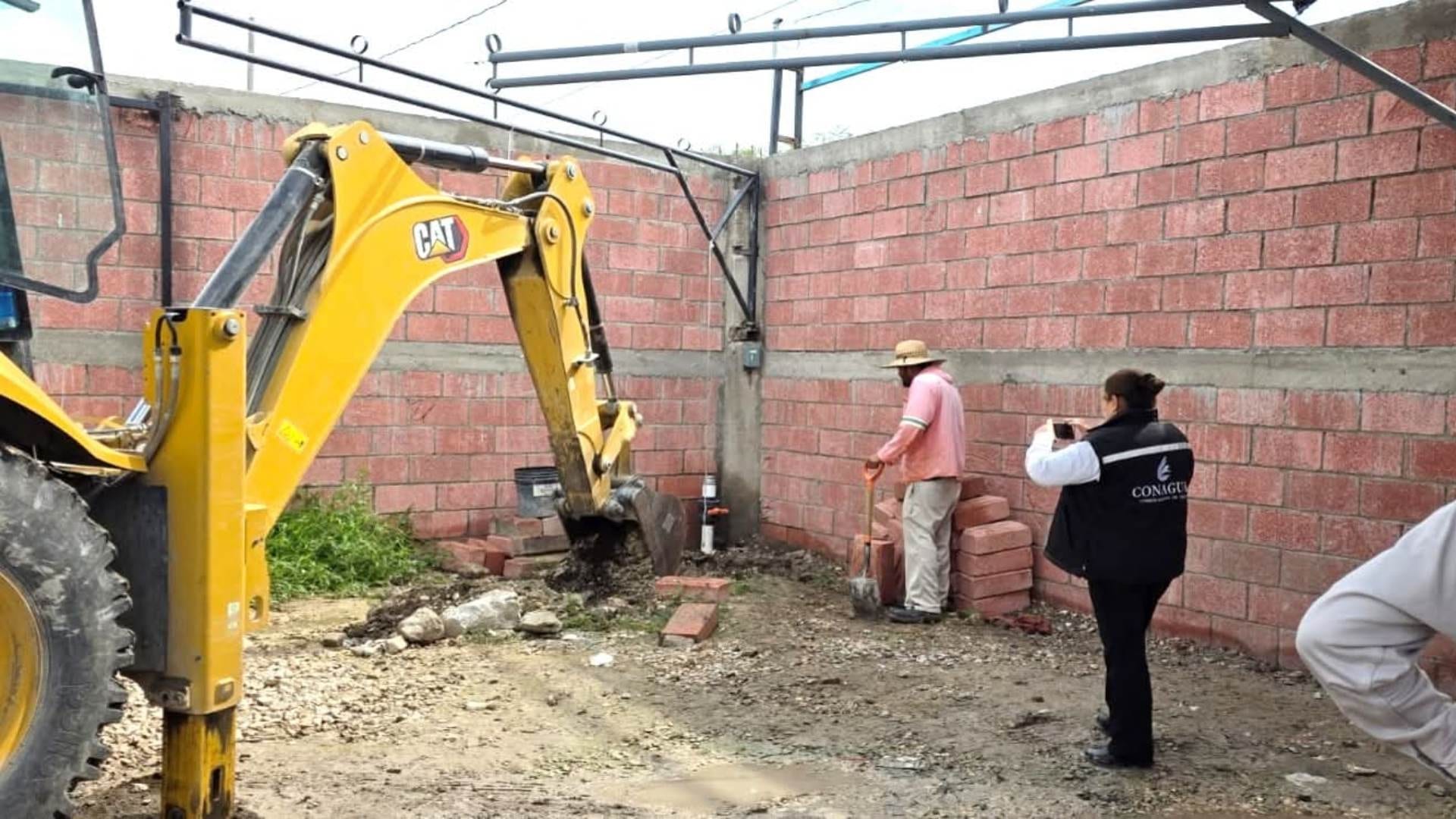 Una funcionaria con chaleco de Conagua fotografía con su celular una excavadora amarilla y a un trabajador con pala, junto a un muro de tabique.
