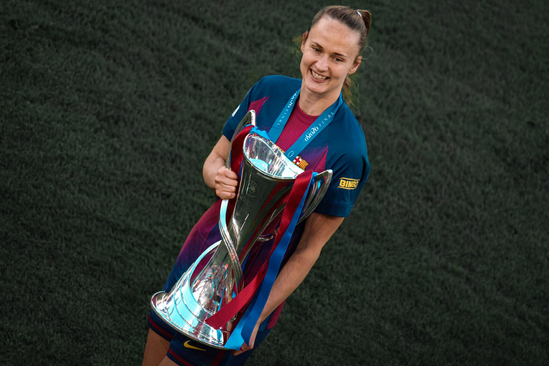 Una jugadora del Barcelona, con una medalla al cuello, sonríe mientras sostiene el trofeo de la Champions League Femenina junto al pecho, sobre el césped.