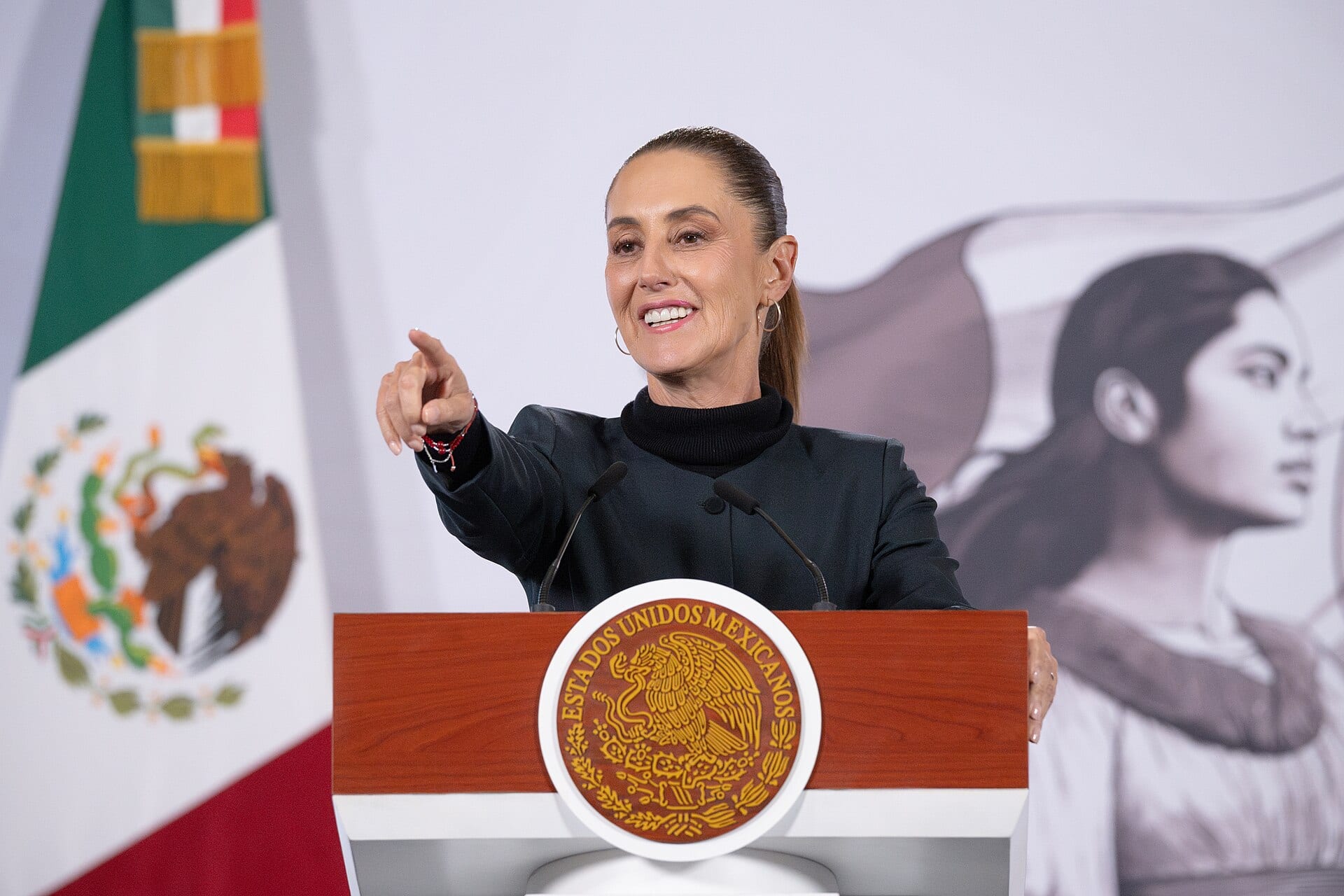 Claudia Sheinbaum sonríe y señala con el dedo desde el podio presidencial, con la bandera de México detrás.