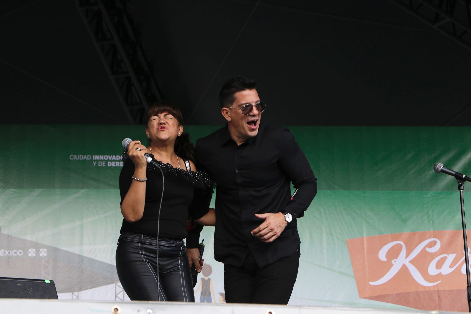 El cantante Yahir, con lentes oscuros, canta junto a una mujer con micrófono en un templete al aire libre, con una manta verde de fondo.