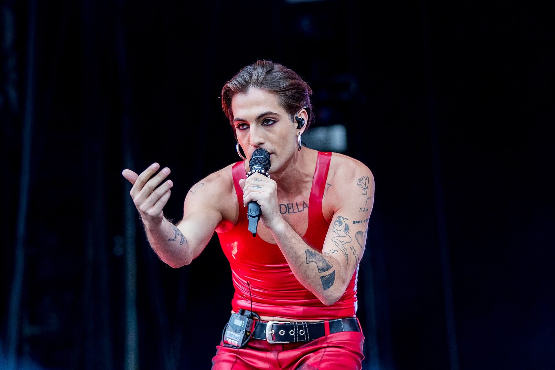 Damiano David canta con un micrófono en la mano, con top rojo y tatuajes visibles en los brazos, sobre un escenario oscuro.