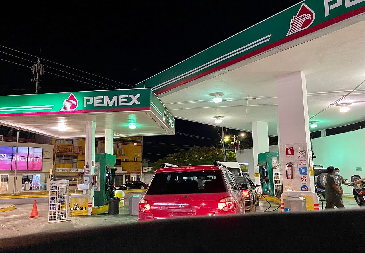 Un automóvil rojo carga combustible de noche bajo el techo iluminado de una gasolinera Pemex.