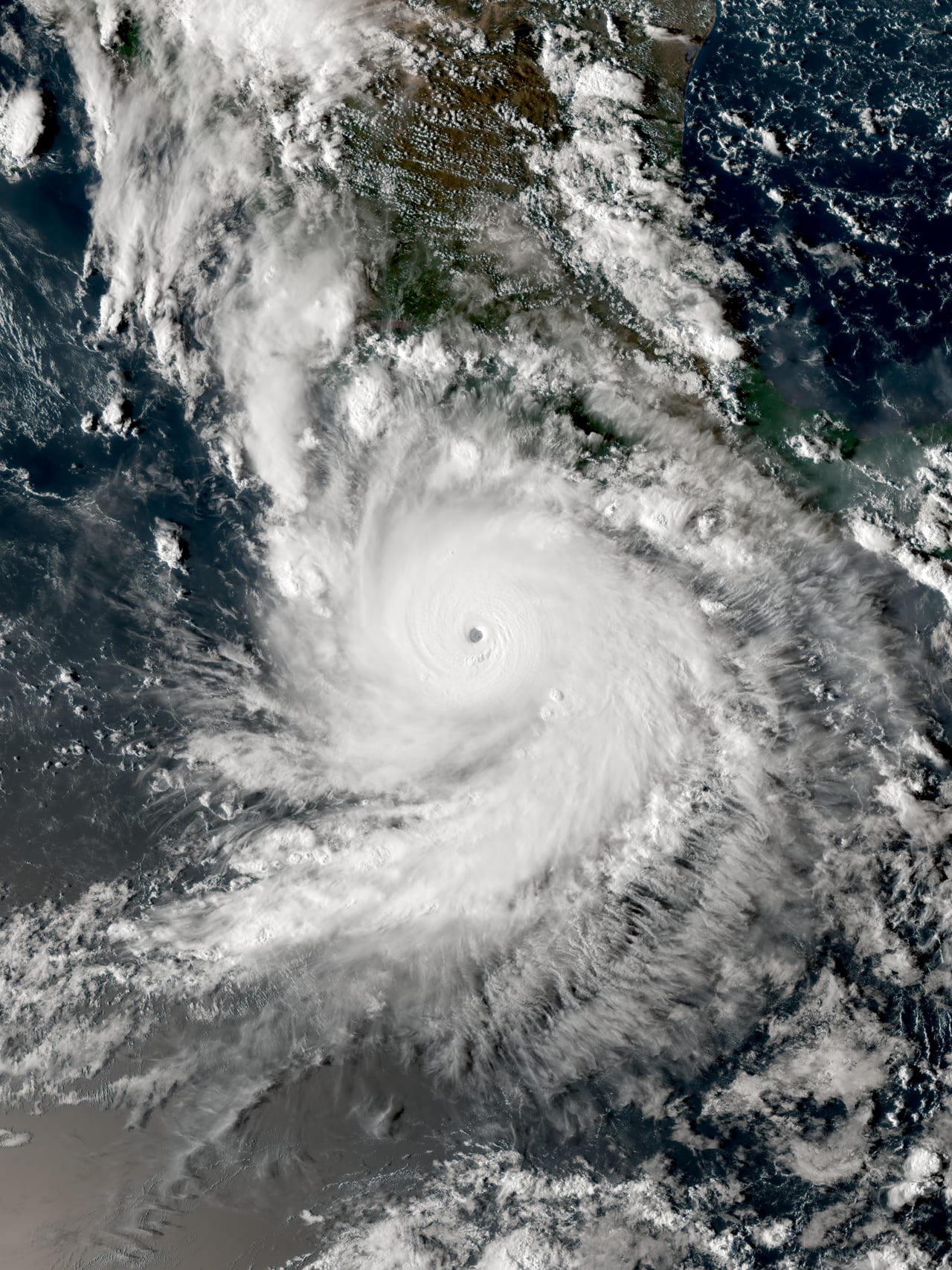 Una masa nubosa blanca y densa gira en espiral sobre el océano, con un ojo bien definido en el centro y una costa continental visible en la esquina superior derecha.