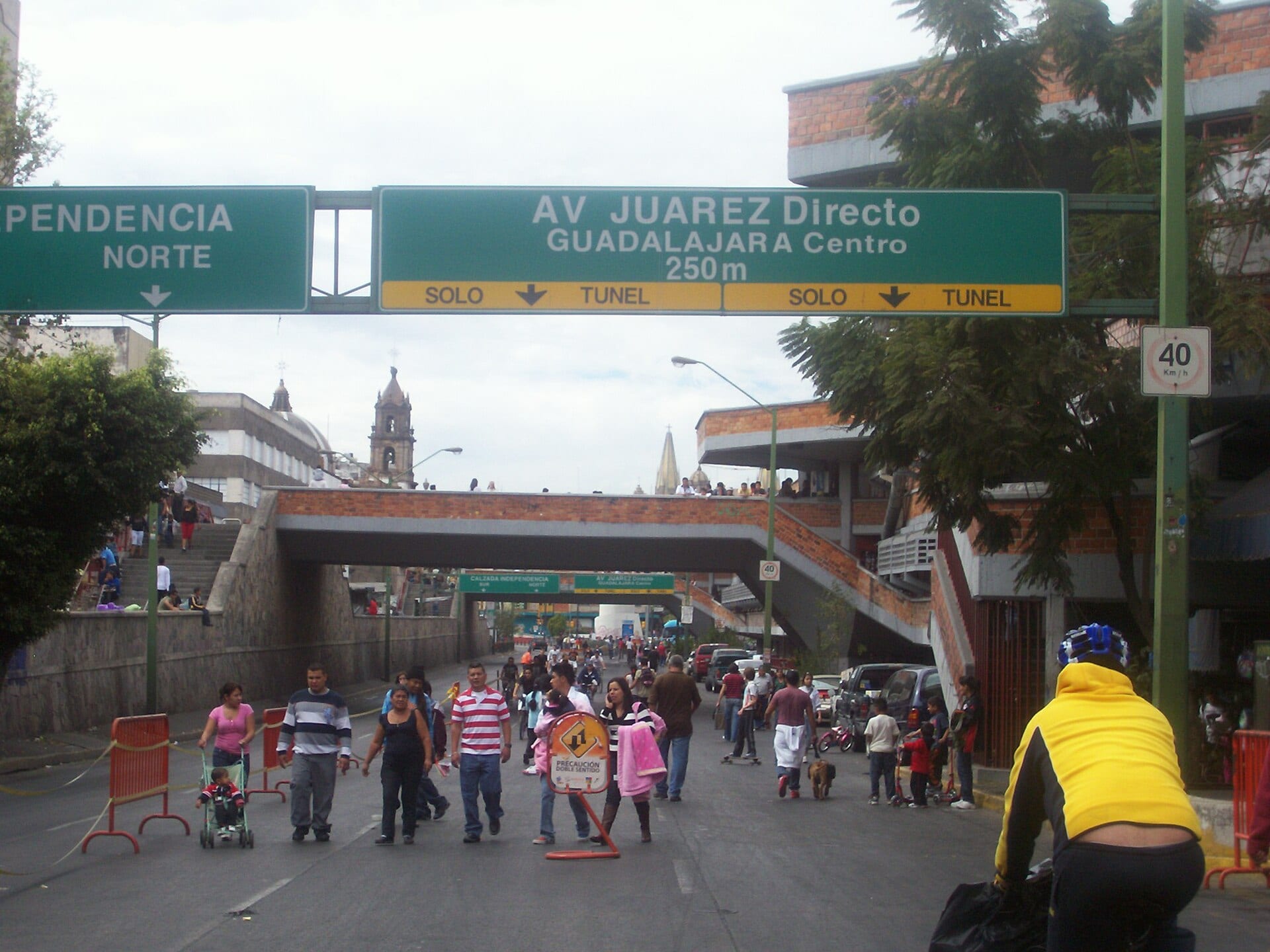 Una calle cerrada al tráfico en el centro de Guadalajara, con peatones caminando bajo señalamientos viales y una torre de iglesia al fondo.