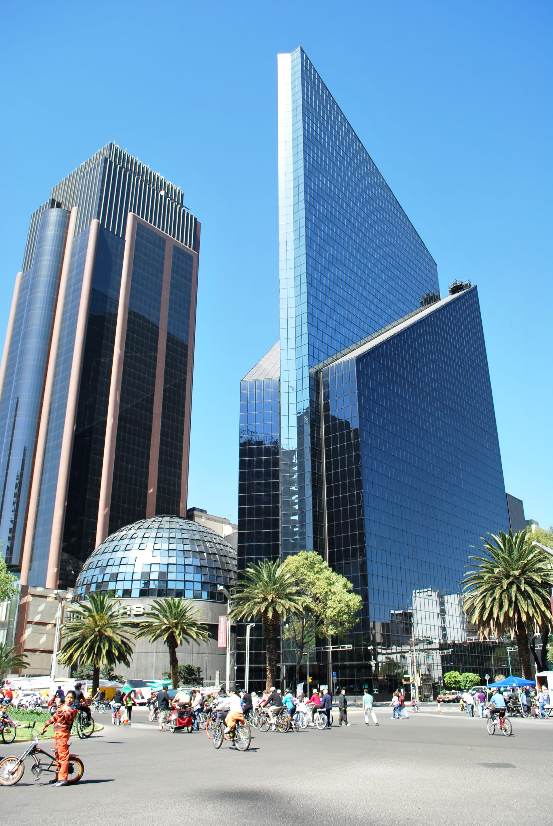 Rascacielos de fachada de vidrio junto a un edificio con cúpula acristalada sobre una avenida con palmeras.