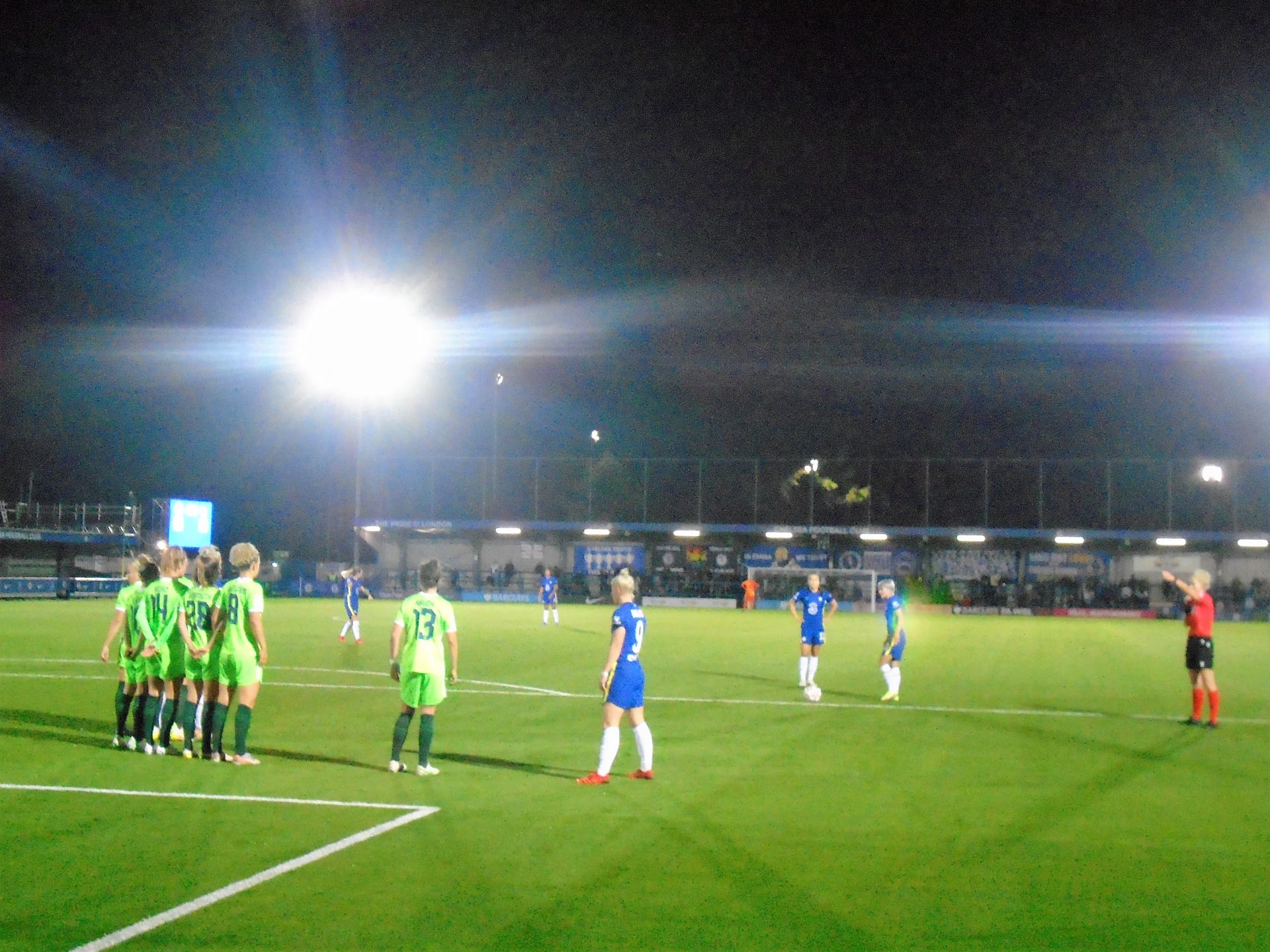 Jugadoras del Chelsea con uniforme azul y del rival con uniforme verde se colocan antes de un saque de esquina bajo las luces del estadio.
