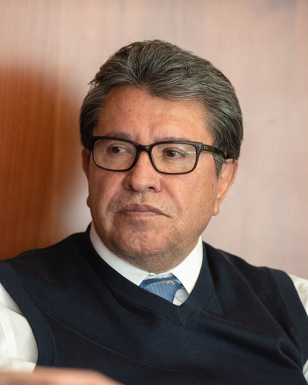 Ricardo Monreal Ávila, con lentes y chaleco azul marino sobre camisa blanca, mira hacia un lado con gesto serio.